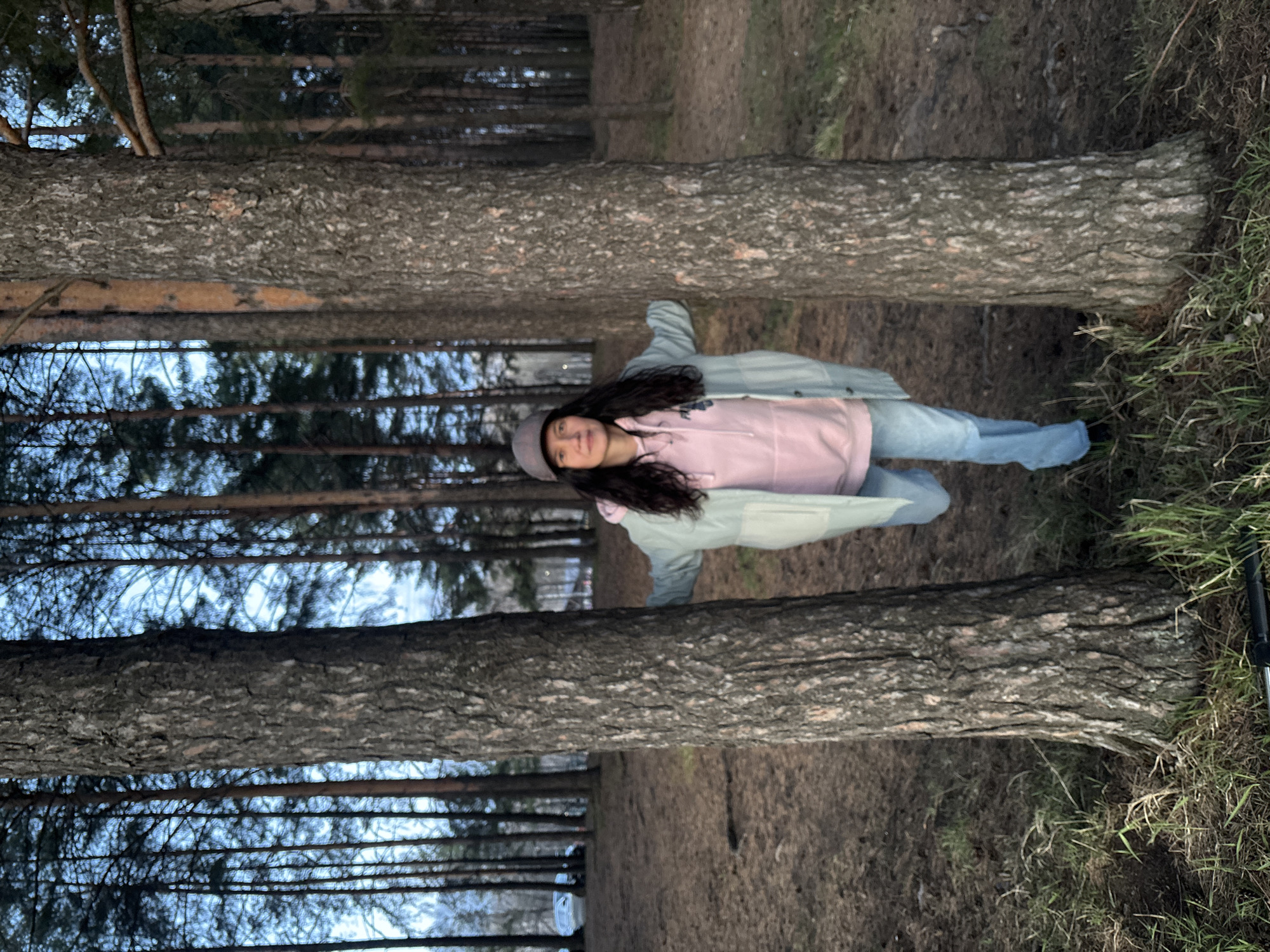 A vertical full-length photograph captures a young woman standing amidst a coniferous forest in what appears to be Chaikovsky, Russia. She is positioned between two prominent, thick tree trunks with rough, textured bark that frame the scene. The woman, with long dark hair, is looking towards the viewer with a gentle smile. She is dressed in a casual outfit consisting of a light pink hoodie, a light blue or grey jacket worn over it, light blue jeans, and a grey cap or beanie.
The setting is a dense wooded area, characterized by numerous tall, slender trees with dark bark and evergreen foliage visible in the background. The ground is a mix of brown dirt, fallen pine needles, and patches of green grass, particularly noticeable on the right side of the frame where a black, pole-like object (possibly a ski or walking stick) is partially visible. The sky, seen through the tree canopy, has a soft, diffused blue hue, suggesting either an overcast day or the soft light of late afternoon or early evening. The overall lighting is subdued, creating a calm and natural atmosphere. No text is visible in the image.