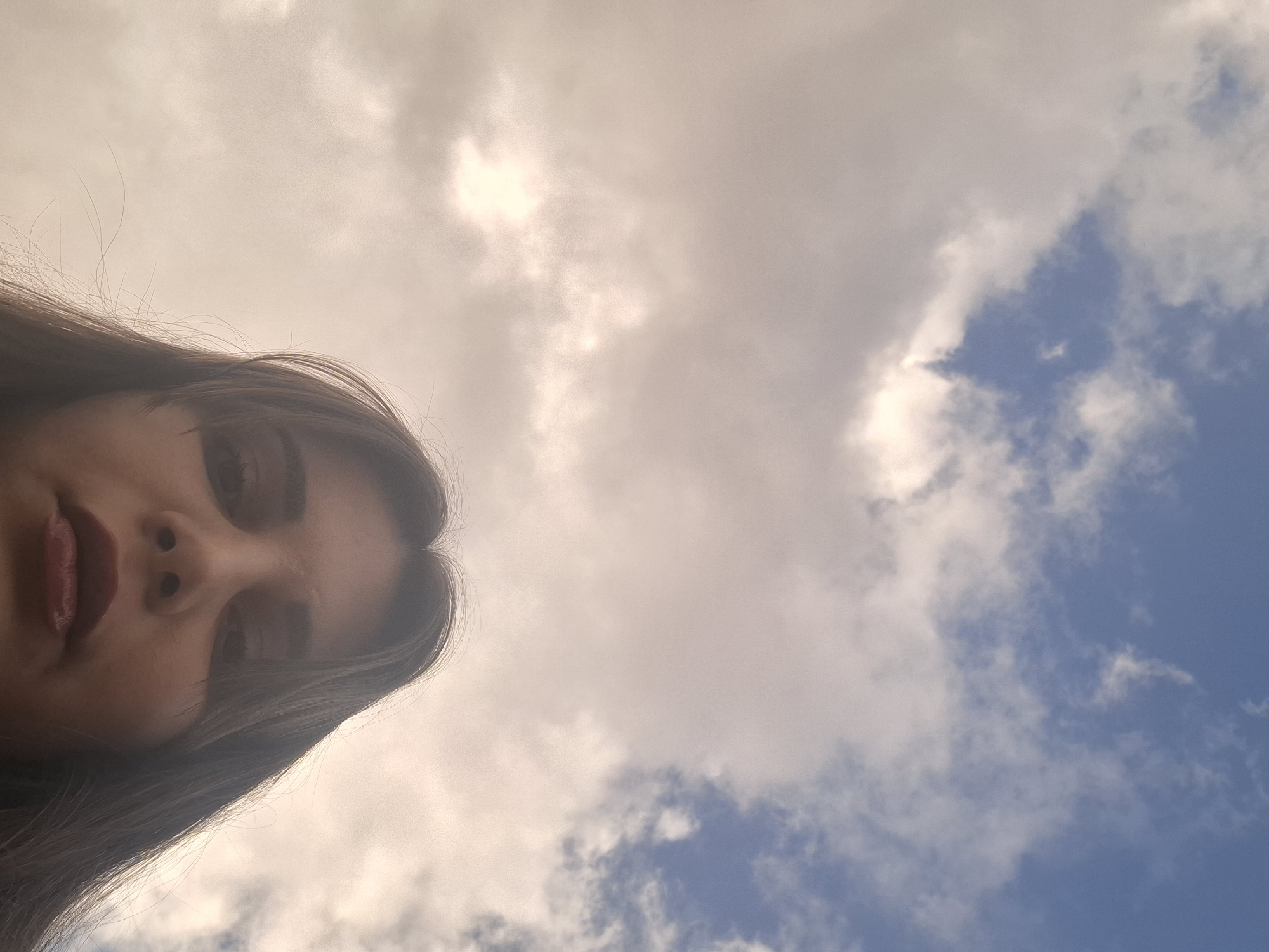 A selfie of a person with long, brown hair with visible red lipstick is in the lower-left corner of the frame. The person's head is tilted to the side. The background is a partly cloudy sky with sections of blue. The location cannot be confirmed.