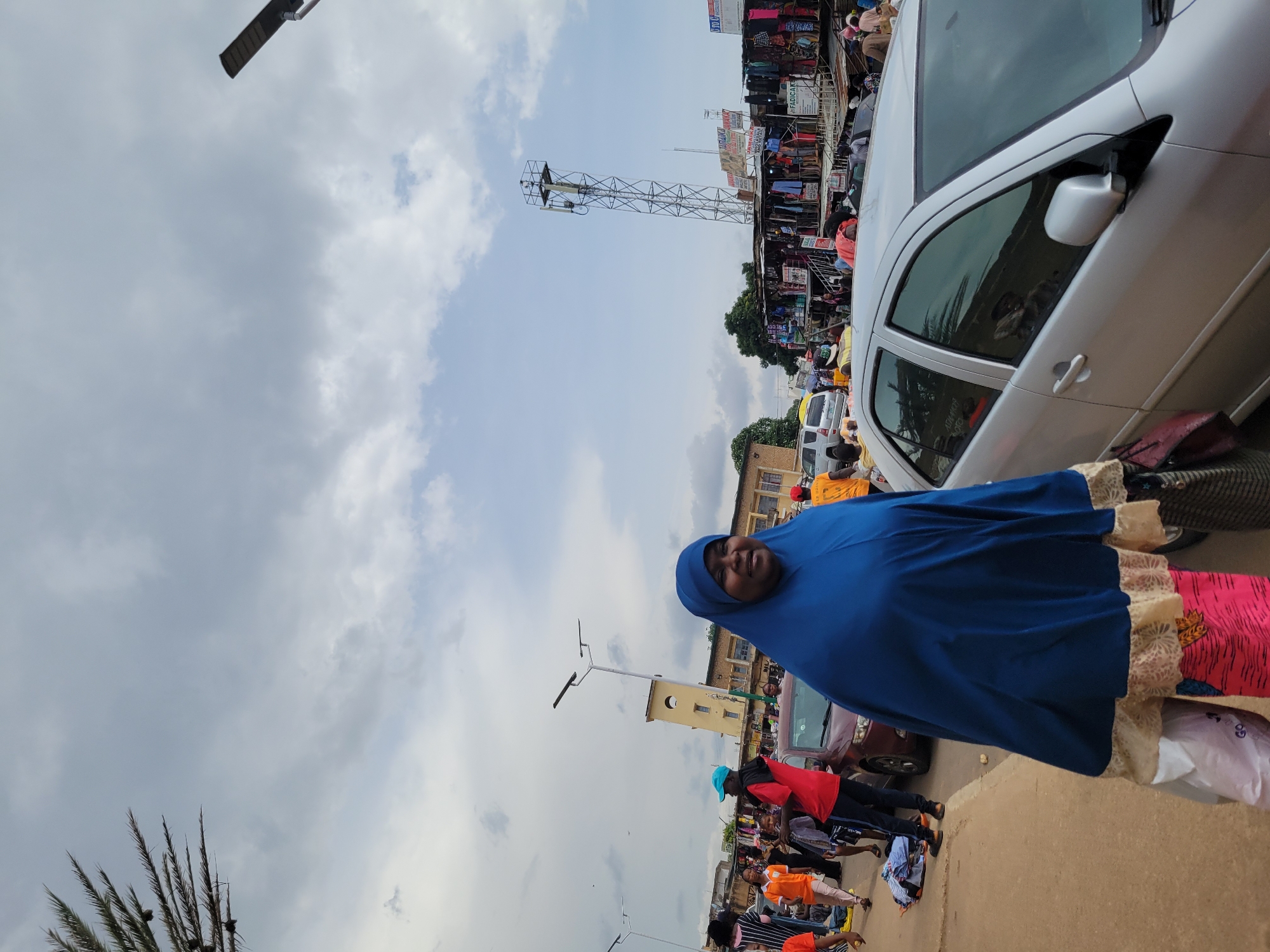 An outdoor street scene shows a woman wearing a blue hijab and a vibrant pink and orange patterned skirt. She is carrying a white shopping bag in her left hand and a patterned bag in her right hand. In the background, several cars are parked or driving, including a silver sedan. Various market stalls are set up along the street, displaying clothing and other goods. People are walking around and browsing the stalls. A cell tower is visible in the distance. The sky is cloudy.