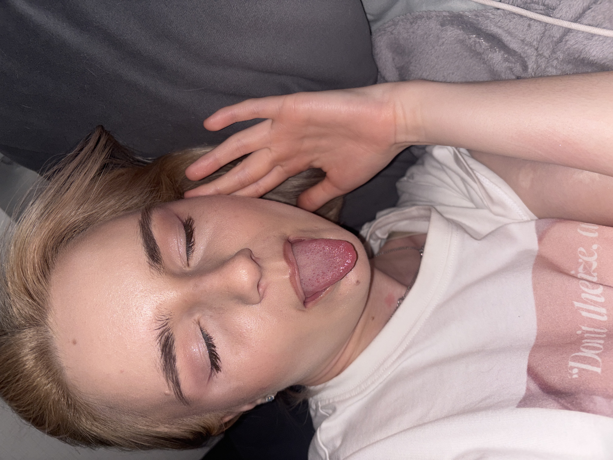 A close-up, high-angle shot captures a person lying down, seemingly resting or sleeping. The person, who appears to be young, has light-colored hair, possibly blonde or light brown, and light skin. Their eyes are closed, and their mouth is slightly open with their tongue partially visible. A small mole is noticeable on their forehead.
The person is wearing a white t-shirt with a pink section on the right side. On the pink part of the shirt, white text is visible, partially reading, "Don't there." A silver chain is around their neck. One of their hands is resting on the back of their head, near their ear, while the other arm is visible lower down, suggesting they are lying on their side.
The background consists of dark grey and lighter grey fabrics, possibly bedding or a couch, which creates a soft, indoor setting. The lighting is somewhat dim, suggesting an indoor environment, possibly at night or in a room with subdued lighting. There are no clear cues about the specific location or time of day beyond the indoor setting. The image evokes a sense of intimacy and vulnerability, given the person's relaxed posture and closed eyes.