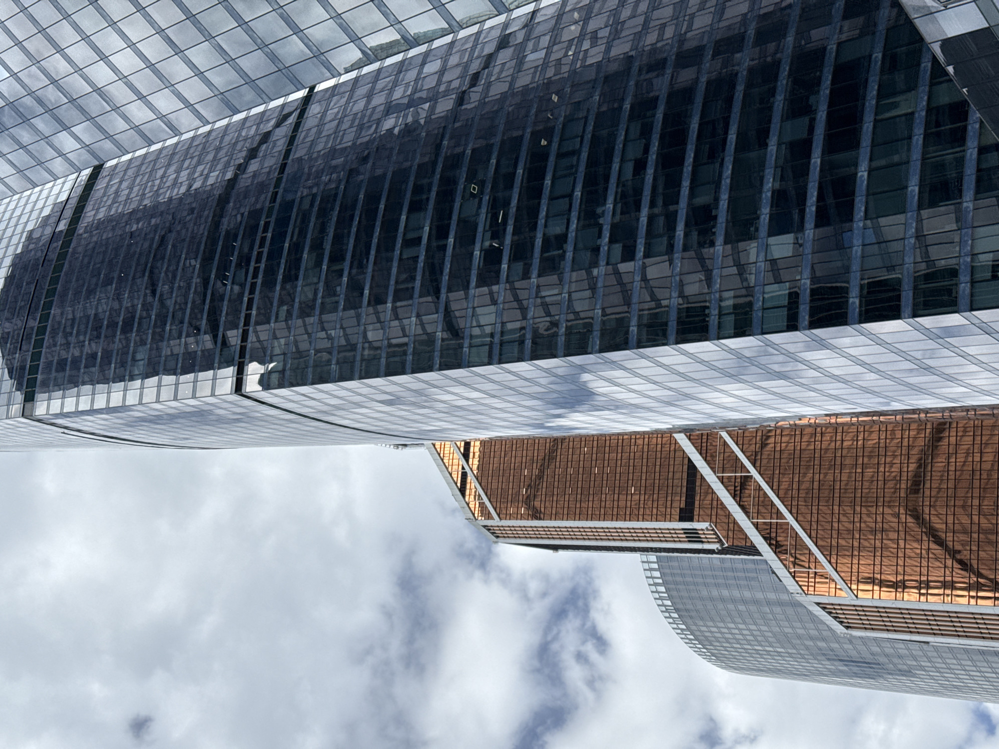 The image is an upward-looking shot of modern skyscrapers. The foreground features a cloudy sky. The bottom skyscraper has a brown, grid-like facade. The top skyscraper is taller and has black glass windows. The tops of the skyscrapers are lined with light grey glass.