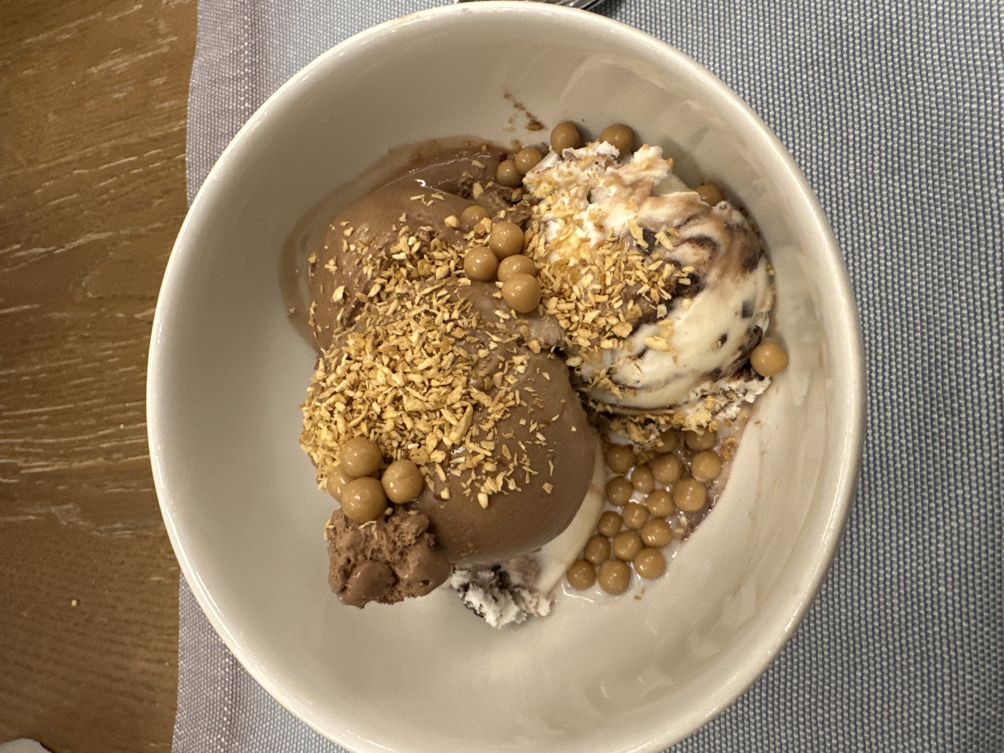 An overhead shot presents a white ceramic bowl filled with scoops of ice cream and various toppings. The bowl contains at least two distinct ice cream flavors: a rich dark brown, likely chocolate, and a lighter, possibly vanilla or cookies and cream flavor with dark swirls.
Both ice cream varieties are generously adorned with golden, flaky pieces, which appear to be toasted coconut or finely chopped nuts. Additionally, numerous small, round, light brown candies or crispy pearls are scattered across the ice cream and within the bowl. Some of these spherical toppings are clustered on the top of the dark brown scoops, while others are submerged in a slightly melted puddle of ice cream towards the bottom right of the bowl.
The white bowl is placed on a light blue-grey placemat with a subtle woven texture. To the left of the placemat, a portion of a wooden table surface with visible grain is captured. The scene suggests an indoor setting, likely a dining area, where a dessert has been prepared or is being served. No individuals or visible text are present in the image.