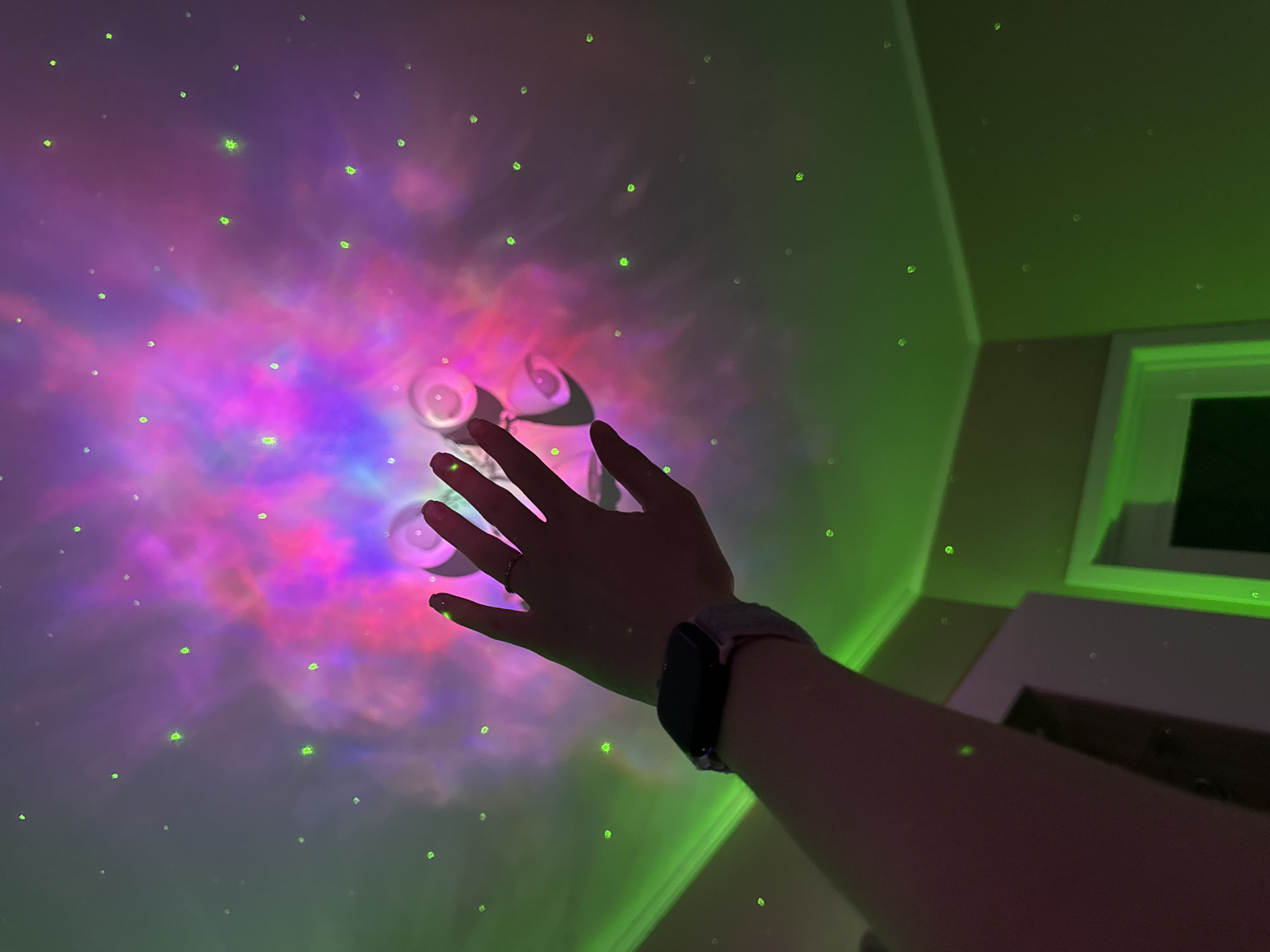An indoor scene is captured from a low angle, looking upwards towards a ceiling and upper walls bathed in vibrant, projected light. A person's left arm extends into the frame from the lower right, with the hand reaching towards the center of the ceiling. A silver-toned ring adorns the fourth finger, and a dark-faced smart watch with a textured light-colored band is worn on the wrist.
The ceiling is illuminated by a galaxy or nebula projector, creating a swirling expanse of purple, pink, and blue hues. Numerous small, bright green laser dots, resembling stars, are scattered across the ceiling and upper walls. A distinct, solid green laser line runs horizontally along the top right corner of the room, highlighting the architectural details.
At the center of the projected nebula, a white projector device is visible, mounted on the ceiling, with two circular elements that appear to be light sources. The room is otherwise dimly lit, allowing the colorful projections to be the dominant feature. A portion of a window frame is visible on the right, catching some of the green light. The overall atmosphere conveyed is one of wonder and immersion in the artificial cosmic environment. No text is visible in the image.