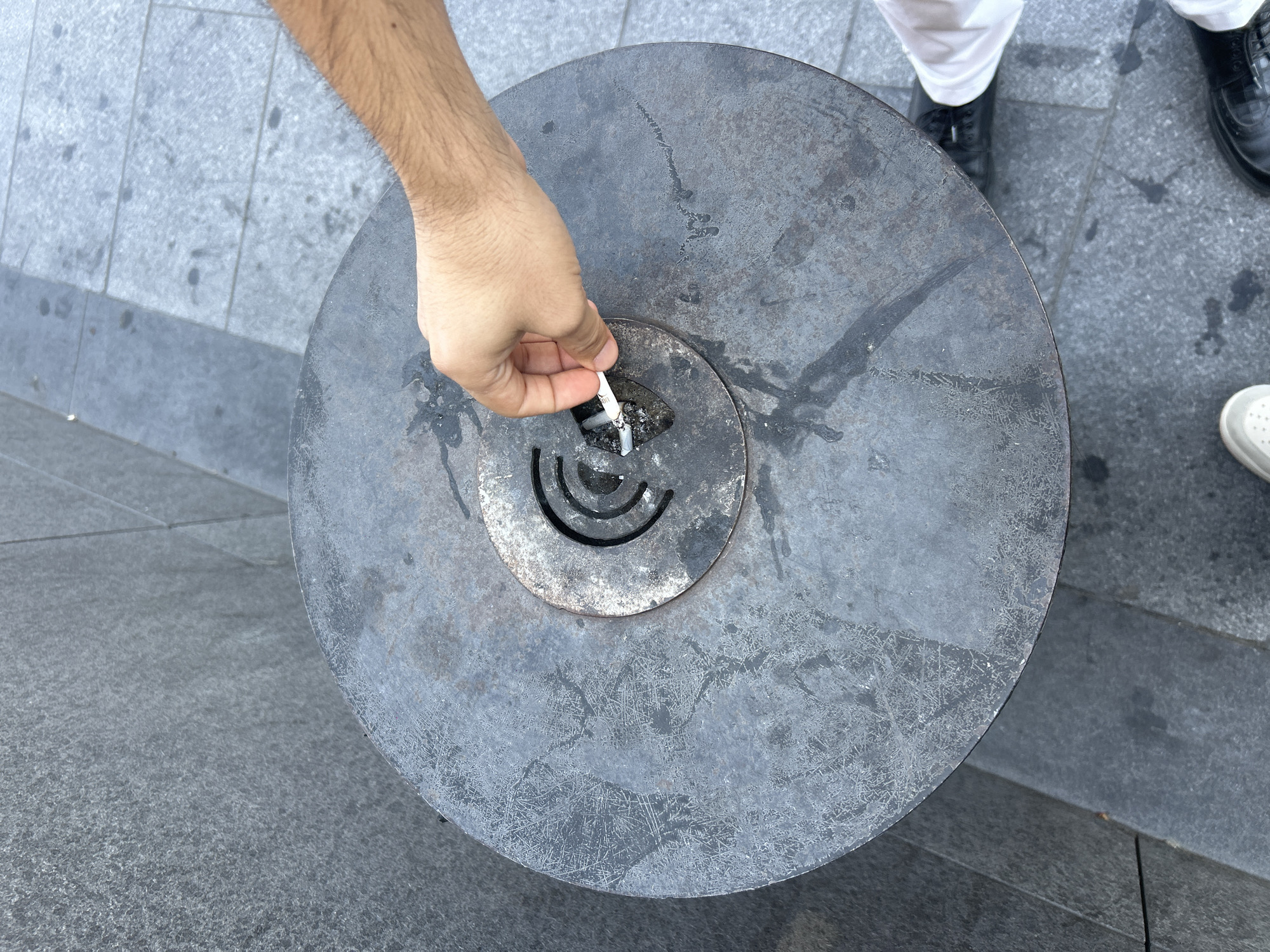 A person's hand is shown holding a lit cigarette, about to extinguish it in a round, metal ash receptacle. The receptacle is situated outdoors on a paved surface, likely in a public area, next to a textured grey wall. The ash receptacle has a central, circular opening with concentric rings. The individual's arm is hairy and tanned, and they are wearing white trousers and black shoes. The pavement is made of grey rectangular tiles. The scene is suggestive of a public space in Baku, Azerbaijan, where smoking is common.