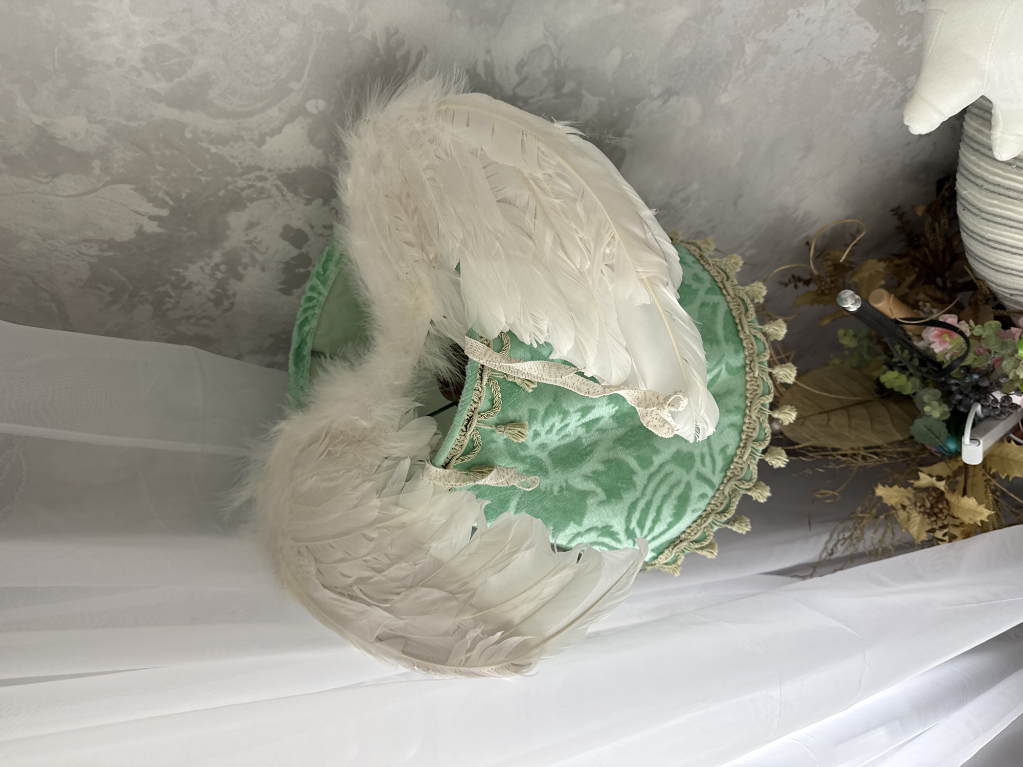 In a studio setting in Unknown, Unknown, a pair of white feather angel wings are draped over a teal, patterned lampshade. The lampshade is adorned with gold trim and small, cream-colored tassels. A delicate lace ribbon is attached to the front of the lampshade. The background features a sheer white curtain on the left and a textured, gray wall on the right. Below the lampshade, a collection of dried flowers and faux greenery is visible.