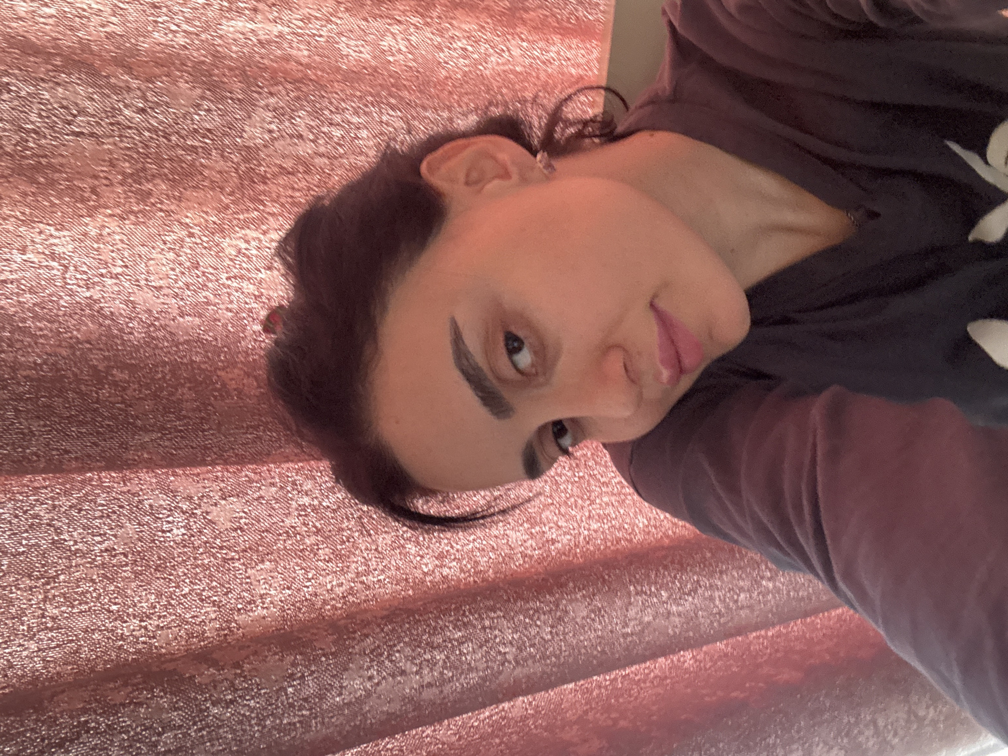 A close-up portrait features a young adult posing against a shimmering, rose-gold or coppery-pink textured fabric background. The person is oriented as if looking upwards and to their right (in a mentally rotated upright view, they are looking slightly to the left of the viewer), captured from the shoulder up. They have dark hair, some strands loosely framing their face, and a visible left ear adorned with a small, reflective earring. Their prominent, dark eyebrows frame dark eyes that look directly towards the camera. Their lips are slightly parted, forming a subtle pout or a thoughtful expression.
They are wearing a dark, possibly black or deep purple, long-sleeved top, with a hint of a white or light-colored pattern, possibly floral, visible on the right side of their chest. The background fabric is richly textured with a woven pattern that catches the light, creating a sparkling effect, and appears to be draped or pleated, forming soft horizontal undulations. The lighting is soft and even, suggesting an indoor setting during the daytime. The overall mood is contemplative or calm. No visible text is present in the image. This photograph was taken in Tbilisi, Georgia.