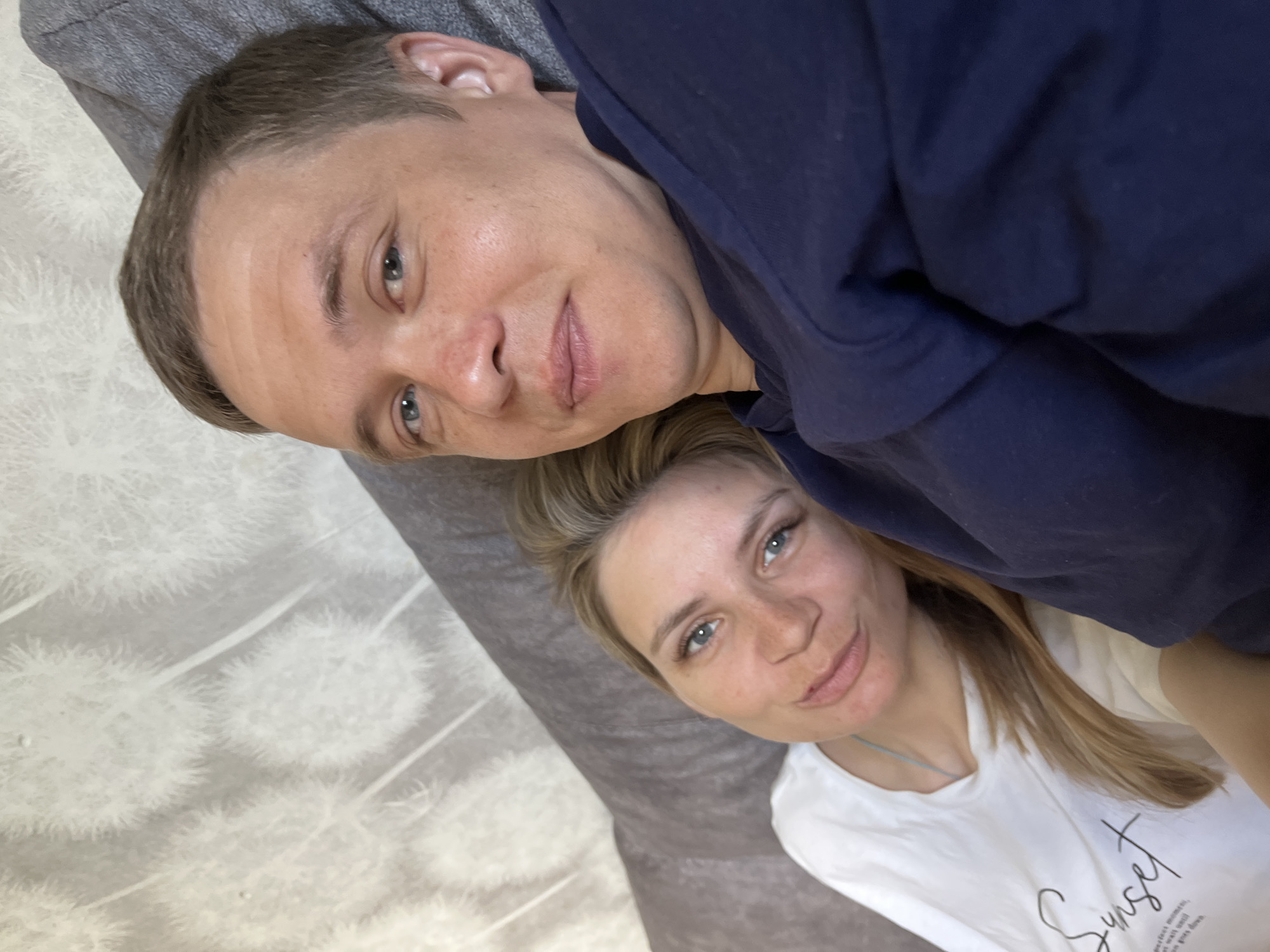 A man and a woman are lying down next to each other, likely indoors. The man, wearing a dark blue shirt, has short brown hair and blue eyes. The woman, wearing a white t-shirt with the word "Sunset" printed on it, has blonde hair and blue eyes. The setting appears to be on a bed with patterned white bedding. The location is likely in Chaikovsky, Russia.