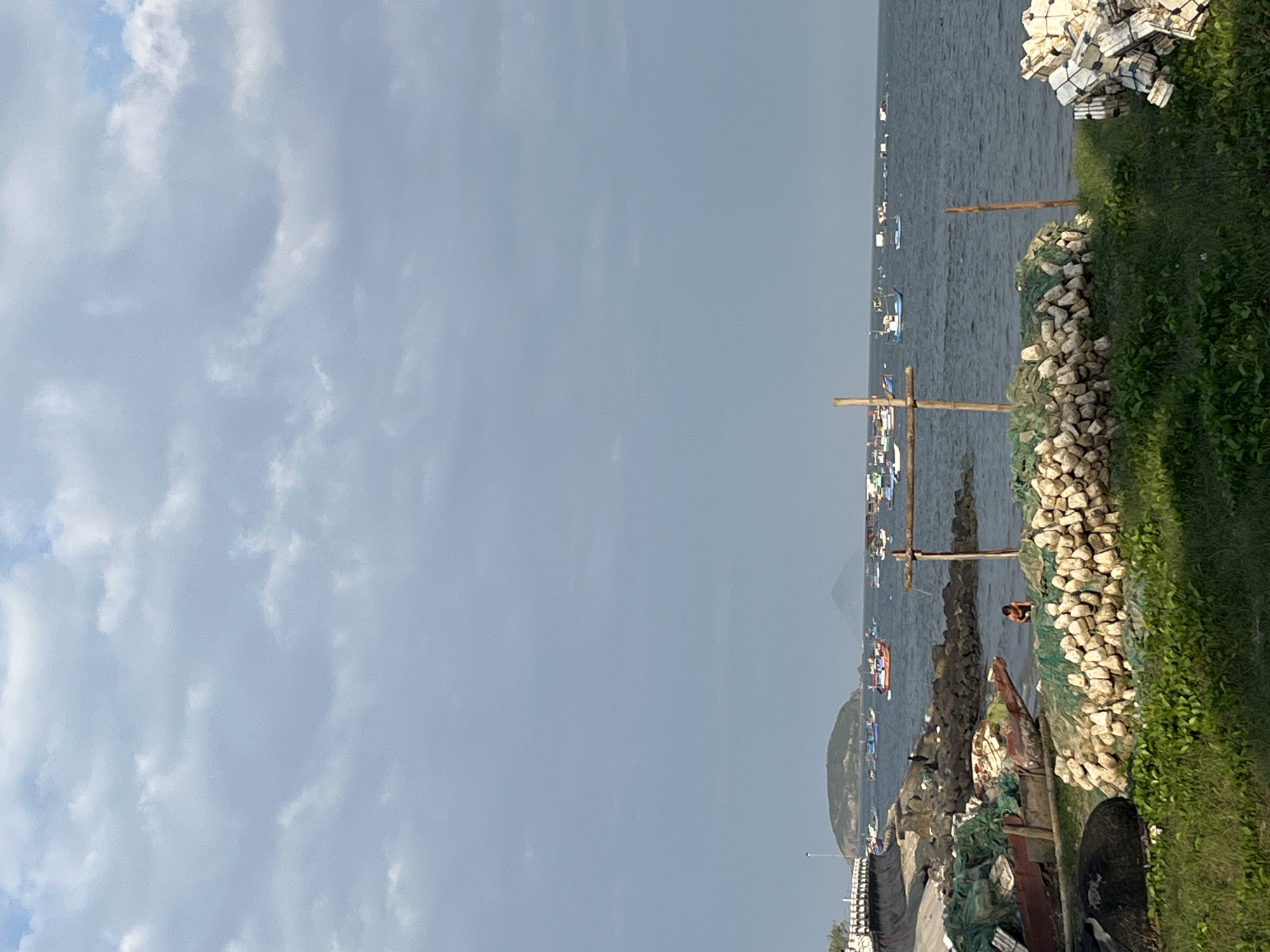 This image depicts a coastal scene in Bac Nha Trang Ward, Vietnam, under an overcast sky featuring light grey clouds with small patches of blue visible in the upper right. The foreground comprises a grassy area transitioning to a rocky shoreline. On the left, a concrete retaining wall with white railings forms a curved structure along the coast. Below this wall, a dilapidated wooden boat, partially covered by green fishing netting, rests on the dark rocks. A single individual, dressed in dark attire, is seated or crouched on the rocky shore near the damaged boat. In the central foreground, a low, rounded mound constructed from numerous small, light-colored stones is visible, topped with green fishing nets. Two wooden poles, arranged in a crude cross shape, are embedded near this mound, with another single pole positioned further right. A large, dark, possibly deflated buoy or tire lies on the grass to the left. To the far right foreground, a stack of white, rectangular bundles is present. The midground features the dark blue-grey bay, populated by numerous small to medium-sized fishing boats of various colors, scattered across the water. In the distant background, two green, hilly landmasses are visible across the horizon.