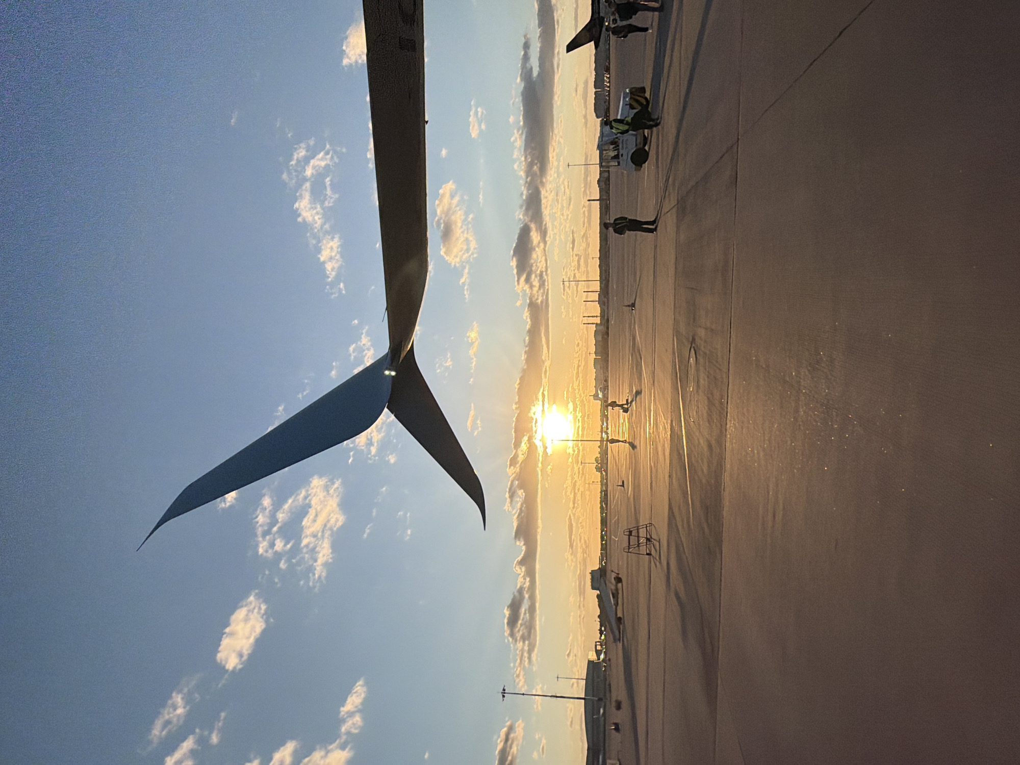 This is a wide-angle, low-angle shot of an airport tarmac at sunset, likely at Vnukovo International Airport (VKO) in Russia. The sun is low on the horizon, casting a warm glow across the scene and creating long shadows.
In the foreground, the edge of an airplane wing is visible, with its distinctive wingtip device extending upwards towards the sky. The wing is dark and casts a shadow across the tarmac.
The tarmac itself is a flat, grey surface, marked with white lines. Scattered across the tarmac are various airport vehicles and equipment, including a tow tractor and a small ladder. Several individuals are present, some appearing to be airport personnel in uniform, while others may be travelers. They are spread out across the tarmac, some standing and others walking.
In the background, more airplanes are visible parked at gates, along with airport buildings and infrastructure. The sky is a clear blue, with a few scattered clouds illuminated by the setting sun. The overall impression is one of a busy airport at the end of a day, with the warm light of sunset creating a serene atmosphere.