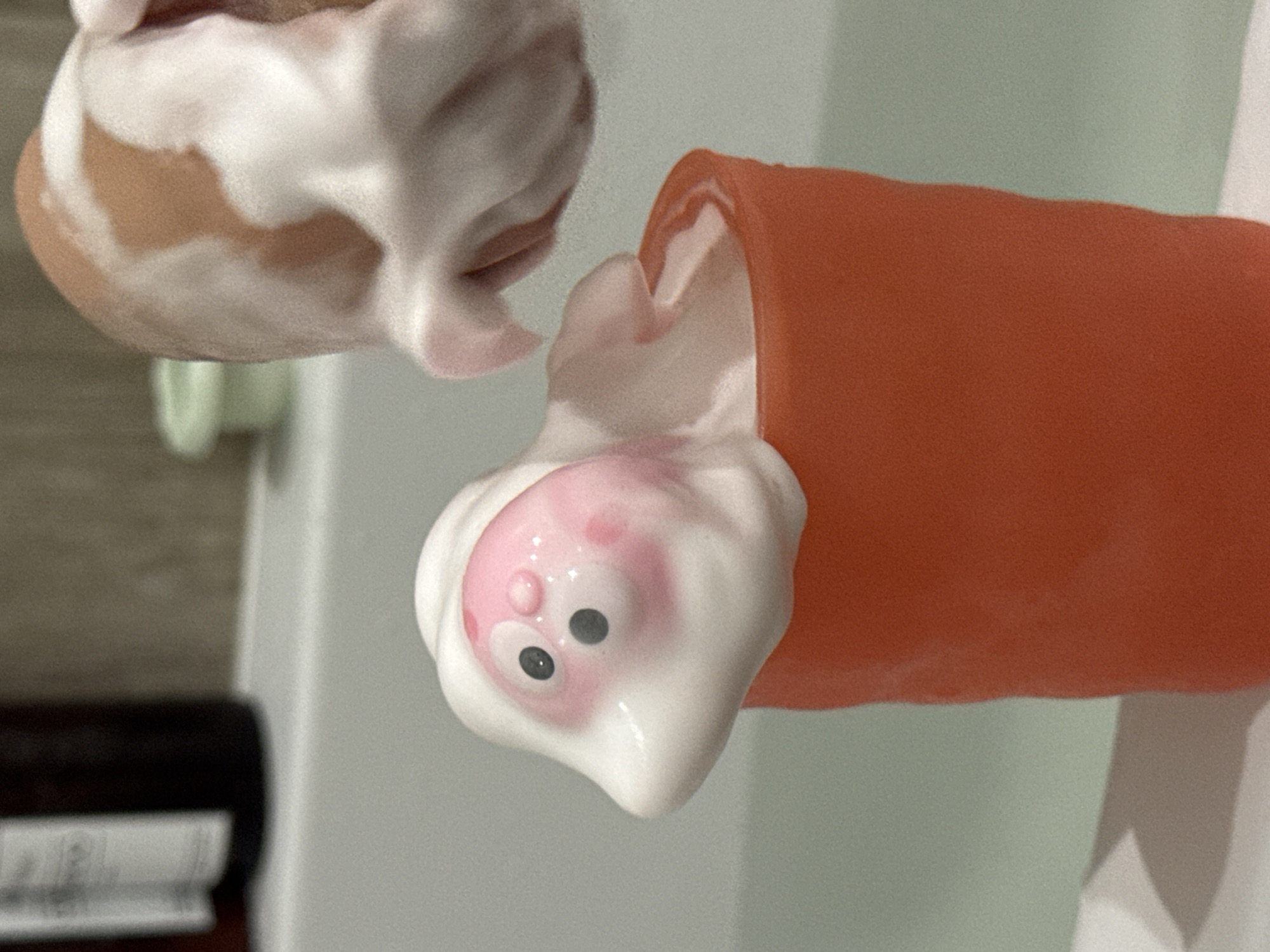 An upside-down close-up shot shows an orange tube with a pink and white character head covered in white foam. Nearby is another object covered in white foam, revealing a tan base underneath. The background consists of a white surface and a dark object on the left. The exact location in the city of Zelenogradsk, Russia is not identifiable from the image.