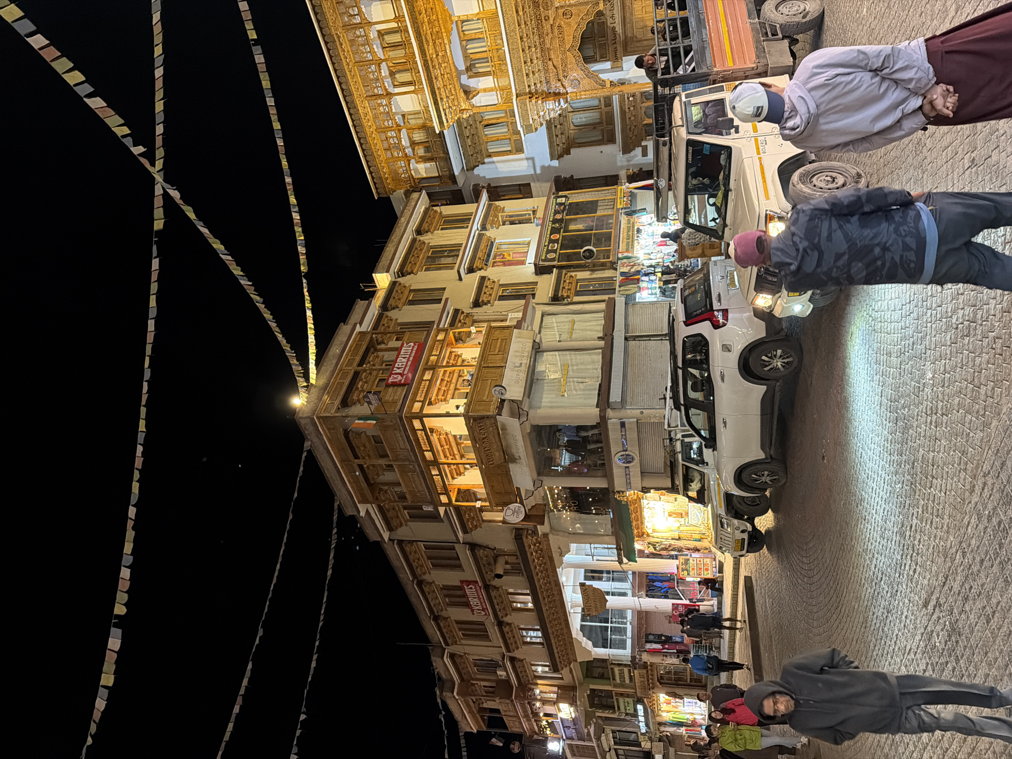 The image depicts a street scene in the city of Leh, India, at night. The street is paved with cobblestones and is illuminated by streetlights and the lights from the buildings. Prayer flags are strung across the street, a common sight in this region.
Several buildings line the street, featuring traditional Ladakhi architecture with intricate woodwork and multiple balconies. These buildings appear to house shops and restaurants, as indicated by signage such as "KARIms," "MENGOON SHOPPING," and "ROYAL BUCKWHEAT RESTAURANT."
In the foreground, two men are standing with their backs to the camera, facing the street. One is wearing a grey hooded sweatshirt and dark pants, while the other is wearing a camouflage jacket and dark pants. Further back, an elderly man in a grey hooded sweatshirt and dark pants walks towards the camera.
Several vehicles are parked on the street, including two white SUVs and a light-colored pickup truck with a flatbed. The pickup truck appears to be carrying passengers and goods in its rear.
Other individuals are visible walking on the sidewalk and in the street, some appearing to be tourists or locals going about their evening. The overall atmosphere suggests a bustling commercial area at night.