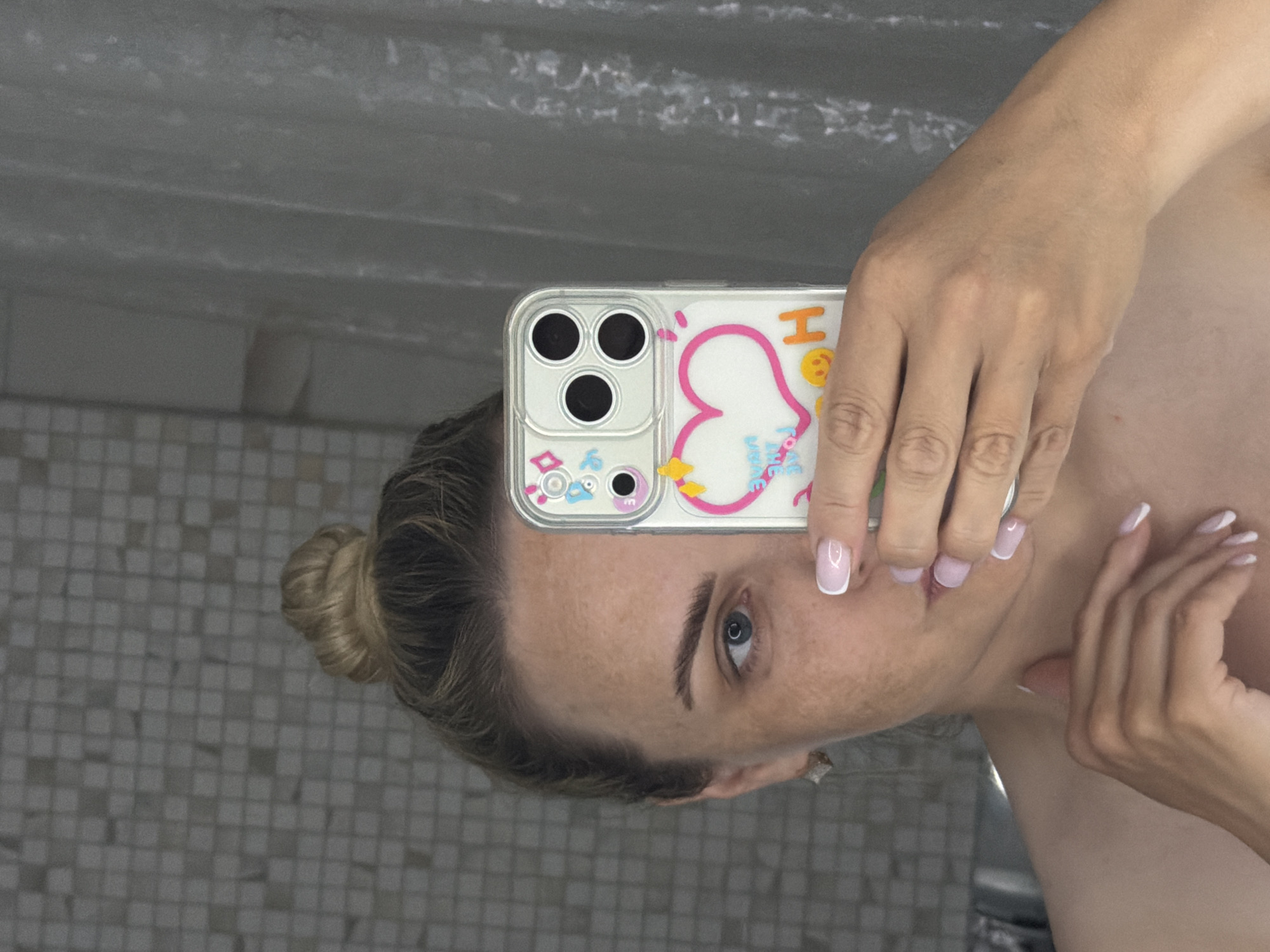 A woman is taking a selfie in a bathroom. The phone is in front of her face. She has light skin, fair hair pulled back into a bun, and blue eyes. Her nails are manicured with white tips. The phone has a clear case with a cartoonish heart design. The bathroom features a tiled wall and a gray countertop. There is no indication of the image being taken in Votkinsk, Russia.