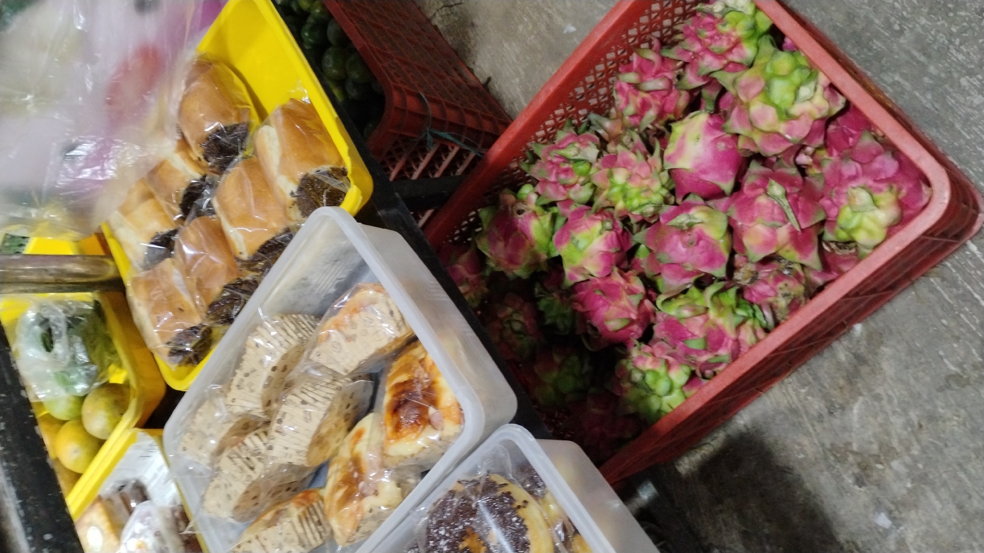 This media file shows a market stall in Banjarsari, Indonesia, filled with various food items. The primary subjects are an assortment of baked goods and dragon fruits. Several loaves of bread, some appearing to be sweet rolls with chocolate filling and others wrapped in patterned paper, are displayed in plastic containers and a yellow tray. Next to the bread, a red basket overflows with vibrant dragon fruits, their pink and green skins a striking contrast. In the background, more fruits, possibly limes or small oranges, are visible in another yellow container, alongside a dark, perforated crate.
The scene appears to be an outdoor market or an indoor market with open stalls. The lighting suggests it could be daytime, with natural light illuminating the products. There are no people directly visible in the frame, but the arrangement of goods implies a vendor has prepared them for sale. The overall impression is one of abundance and freshness, characteristic of a local food market. The ground beneath the displays is a rough, gray surface, possibly concrete or stone. No visible text is present in the image.
