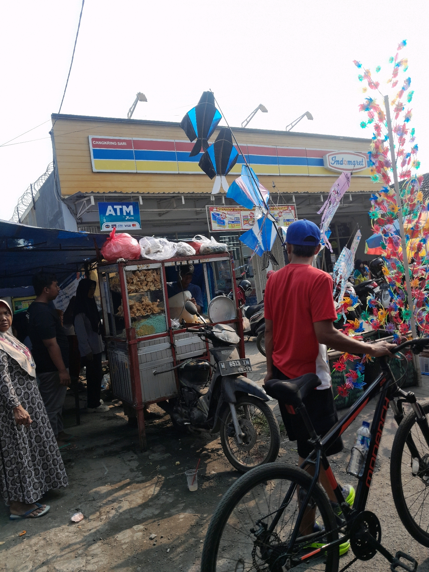 A medium shot captures an outdoor scene in Jogosatru, Indonesia, with a bright, overcast sky. In the background, a yellow-and-red striped awning of an Indomaret convenience store spans the width of the frame, with a prominent "Indomaret" logo on the right. A sign above reads "CANGKRING SARI." A blue ATM sign is visible below the awning.
In the midground, a red food cart with a metal frame displays various fried snacks and noodle products under glass. A person in a blue t-shirt and black shorts stands facing away from the camera, holding onto the handlebars of a black bicycle. The bicycle has a water bottle attached to its frame. Behind the food cart, several motorcycles are parked.
To the left of the food cart, a woman in a patterned dress and headscarf stands facing the camera, her left hand resting on her hip. Next to her, a man in a black t-shirt stands facing the food cart. Another person in a grey shirt and headscarf is also visible near the food cart.
Decorations are scattered throughout the scene. Several black and blue kites, along with pink and blue kites, are suspended from wires above the food cart and store. A colorful display of decorative pinwheels or small windmills, made of brightly colored plastic, stands on the right side of the frame.
The ground appears to be a mix of dirt and asphalt, with some scattered debris and shadows cast by the overhead structures and decorations. A clear plastic cup with a red straw is visible on the ground in the lower right quadrant. The overall impression is a bustling street-side market area adjacent to a convenience store.