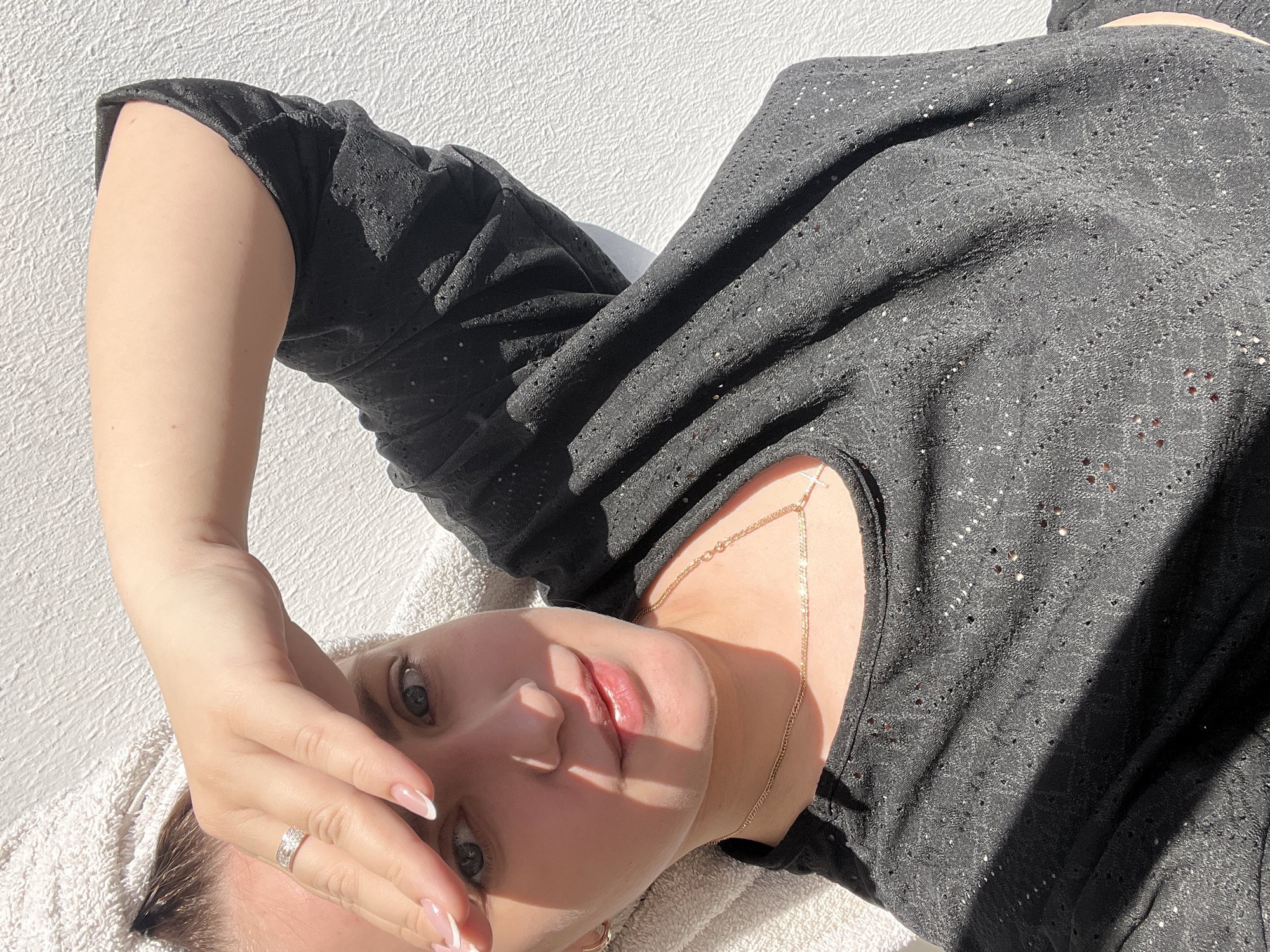 A light-skinned woman is lying on a white towel. Her face is the main focal point. Her right hand is raised to her forehead, with a ring visible on her finger. She is wearing a black patterned dress and a gold body chain with a cross. The background consists of a white wall with a textured surface. The image is taken in an outdoor setting, and the location is Марковский, Russia.