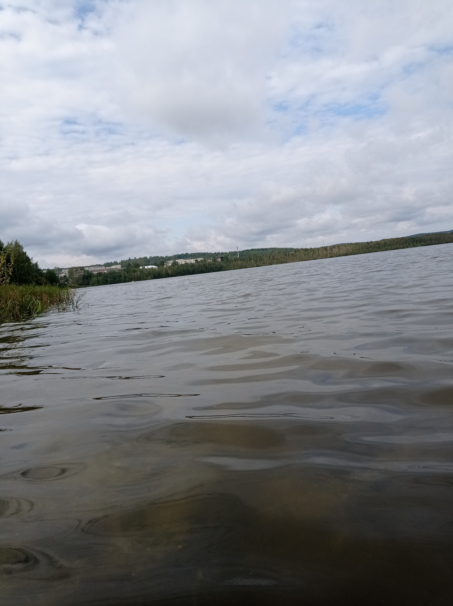 The image shows a wide, calm body of water, likely a lake or river, with a gentle ripple on the surface. The water is dark and reflects the overcast sky and surrounding landscape. On the far bank, there is a line of trees, interspersed with buildings of various sizes, suggesting a settlement or town. The sky is filled with clouds, with patches of blue sky visible. The foreground of the image is dominated by the water, with some vegetation visible on the left bank, including tall reeds. The scene appears to be a natural landscape with human development in the distance. Given the context of Degtyarsk, Russia, this is likely a view of the Iset River or one of the local reservoirs.