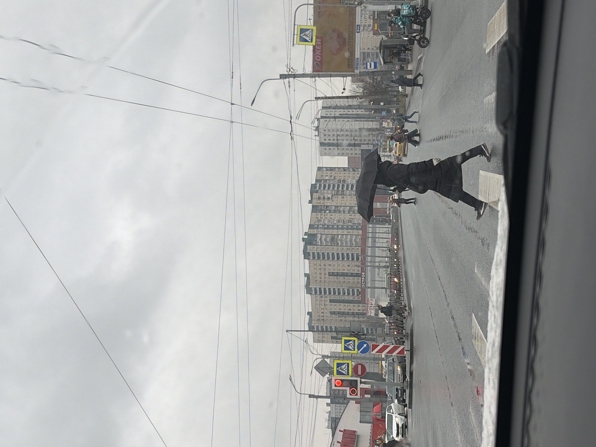 A street scene in Saint Petersburg, Russia, captured on an overcast day with rain. The perspective is from inside a vehicle, likely a car or bus.
In the foreground, pedestrians are crossing the street at a crosswalk. One pedestrian has an umbrella, struggling against the wind. Other pedestrians are also visible.
The street is wet, reflecting the ambient light. Several vehicles are present, including cars, a truck, and motorcycles. Traffic signals are red.
In the background, high-rise apartment buildings dominate the skyline. Street lights and overhead wires run along the road. Signage is visible, including traffic signs with pedestrian crossings. A large billboard displays the word "OKEI".
