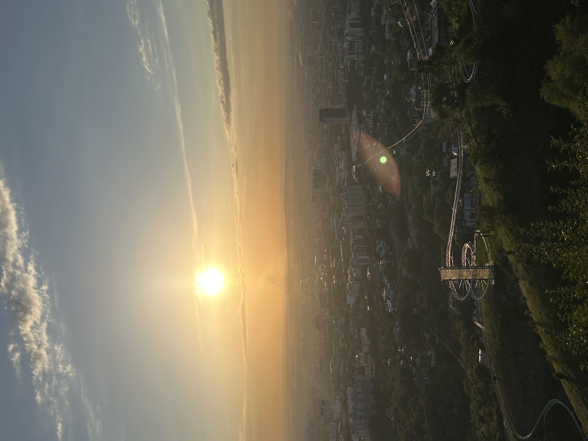 The image captures a panoramic view of Almaty, Kazakhstan, during sunset. The sun is a bright orb in the sky, casting a warm, golden glow over the city. Wispy clouds streak across the pale blue sky. In the foreground, a winding roller coaster track, illuminated by the setting sun, snakes down a grassy hillside. Beyond the roller coaster, the city sprawls out with buildings of varying heights and architectural styles, nestled amongst lush green trees. In the distance, the hazy silhouette of mountains can be seen, adding depth to the cityscape. Smoke rises from industrial chimneys in the far distance, indicating urban activity. A lens flare, a bright green circle, is visible in the mid-ground, obscuring a portion of the city and the roller coaster. The overall scene evokes a sense of tranquility and urban beauty at dusk.