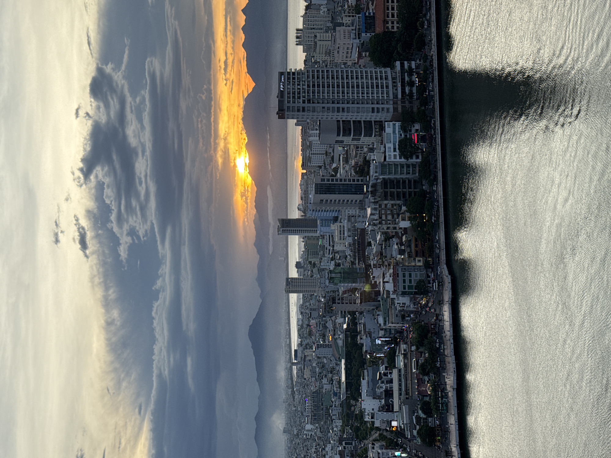 This is an aerial, landscape-oriented photograph taken from a high vantage point, likely a building, overlooking a coastal city at sunset. The sun is low on the horizon, casting a warm, golden glow on the sky and mountains in the distance. Dramatic clouds, tinged with orange and yellow, fill the left side of the frame, suggesting a dynamic weather system.
The scene is dominated by a densely packed urban landscape on the right, with numerous buildings of varying heights, including several tall skyscrapers. The city stretches along a waterfront, with a body of water, possibly a river or the sea, occupying the far right portion of the image. The water's surface is rippled, reflecting the bright light of the setting sun, creating a shimmering effect. A dark shadow, cast by an unseen object or the curvature of the land, creates a stark contrast on the water's surface.
The city appears to be bustling, with lights visible on some buildings and along the streets, indicating the onset of evening. Trees line the waterfront in some areas, providing a touch of greenery against the urban sprawl. The mountains in the background are silhouetted against the bright sky, adding depth to the scene.
There is visible text on one of the buildings: "FPT Camera". The overall mood of the image is serene yet awe-inspiring, capturing the beauty of a sunset over a vibrant city. The time of day is clearly sunset, and the weather appears to be partly cloudy. The location context provided places this scene in Đà Nẵng, Vietnam.