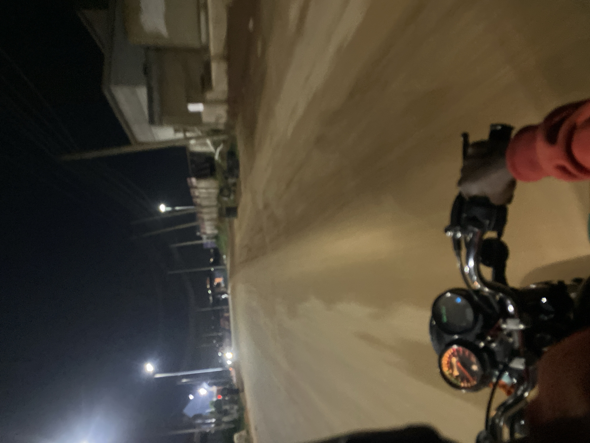 The image captures a first-person perspective from a motorcycle rider at night, with the camera oriented on its side (rotated approximately 90 degrees clockwise). The scene depicts a wide, seemingly unpaved or dusty road stretching into the distance, illuminated by streetlights.
On the left side of the frame, which would be the horizon if the image were properly oriented, are several bright streetlights casting light onto the surrounding area. Beyond the streetlights, outlines of buildings and power lines are visible against the dark night sky. The streetlights appear to be modern, pole-mounted fixtures.
In the foreground, on the right side of the image, parts of the motorcycle are clearly visible. These include the chrome handlebars, the instrument cluster, and a human hand in an orange sleeve gripping the right handlebar. The motorcycle's instrument cluster features at least two gauges; the lower one, likely a speedometer or tachometer, has an illuminated dial with a needle indicating a value, though specific numbers are indistinct. A blue indicator light is visible on the upper part of the instrument cluster.
The overall setting suggests an urban or semi-urban environment in Abuja, Nigeria, characterized by night driving conditions. The road appears relatively clear of other vehicles immediately in front of the motorcycle, though distant lights hint at other traffic or activity further down the road. The dark sky and bright artificial lights confirm it is nighttime. No specific weather conditions are discernible other than a clear sky. There is no clearly readable text in the image.