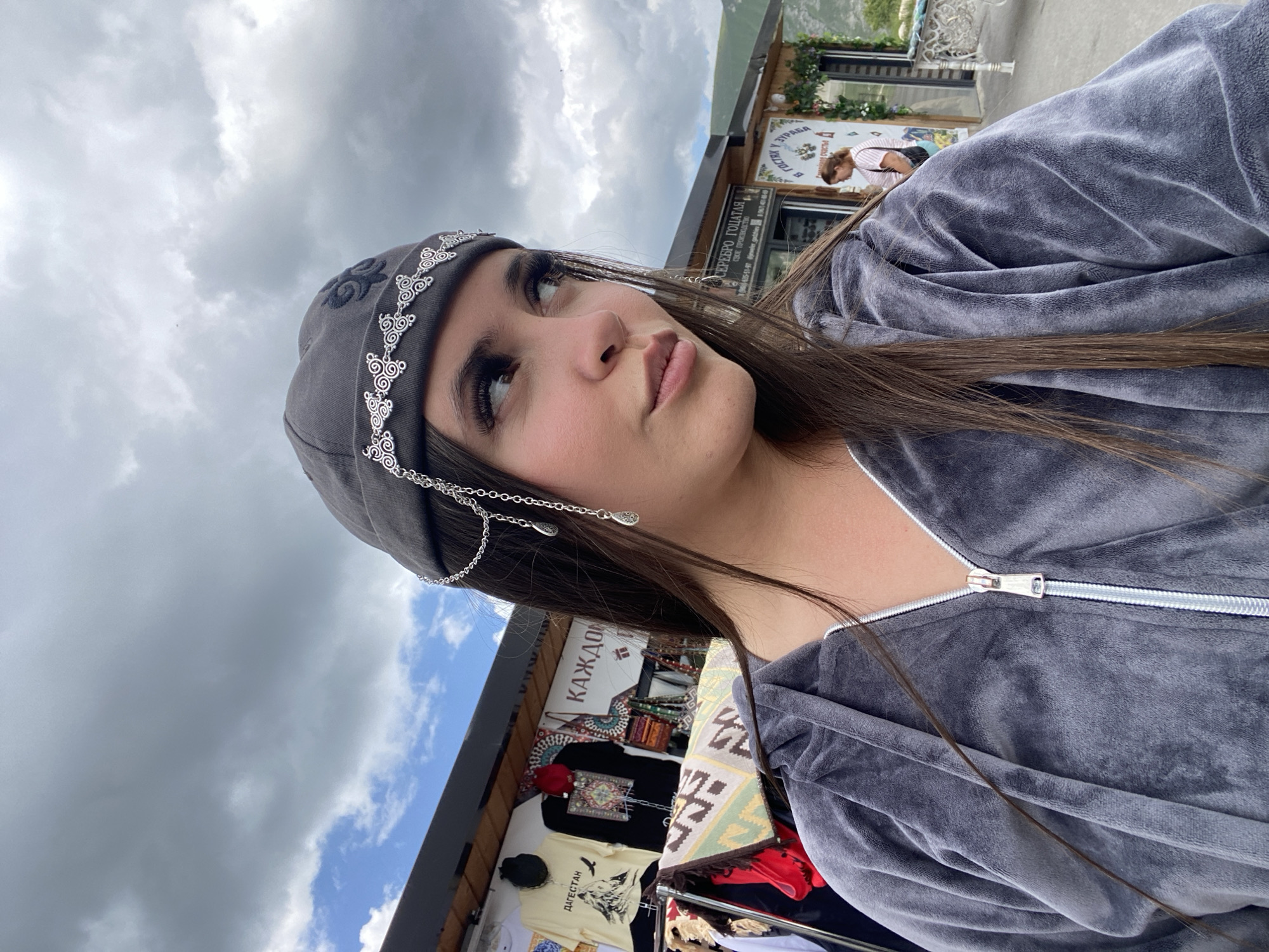 A young woman with long, dark hair and dramatic false eyelashes stands outdoors, looking to her right. She is wearing a grey velour hooded jacket zipped up to her neck and a grey cap adorned with a silver filigree band and dangling chains with teardrop charms. Behind her, a market stall displays various items, including t-shirts with mountainous imagery and colorful fabrics. Signs above the stall are in Russian. Further in the background, a building with Russian signage, including "В ГОСТЯХ У ГУРМА" (Guest of the Gourmet) and "СЕРЕБРО ГОЛЧАТАЯ" (Golchataya Silver), is visible. A woman in a pink and white striped shirt is seen near the entrance of the building. The sky is cloudy. The overall scene suggests a market or roadside attraction in a mountainous region, possibly in or near Tsalkita, Russia, given the Russian language and signage.