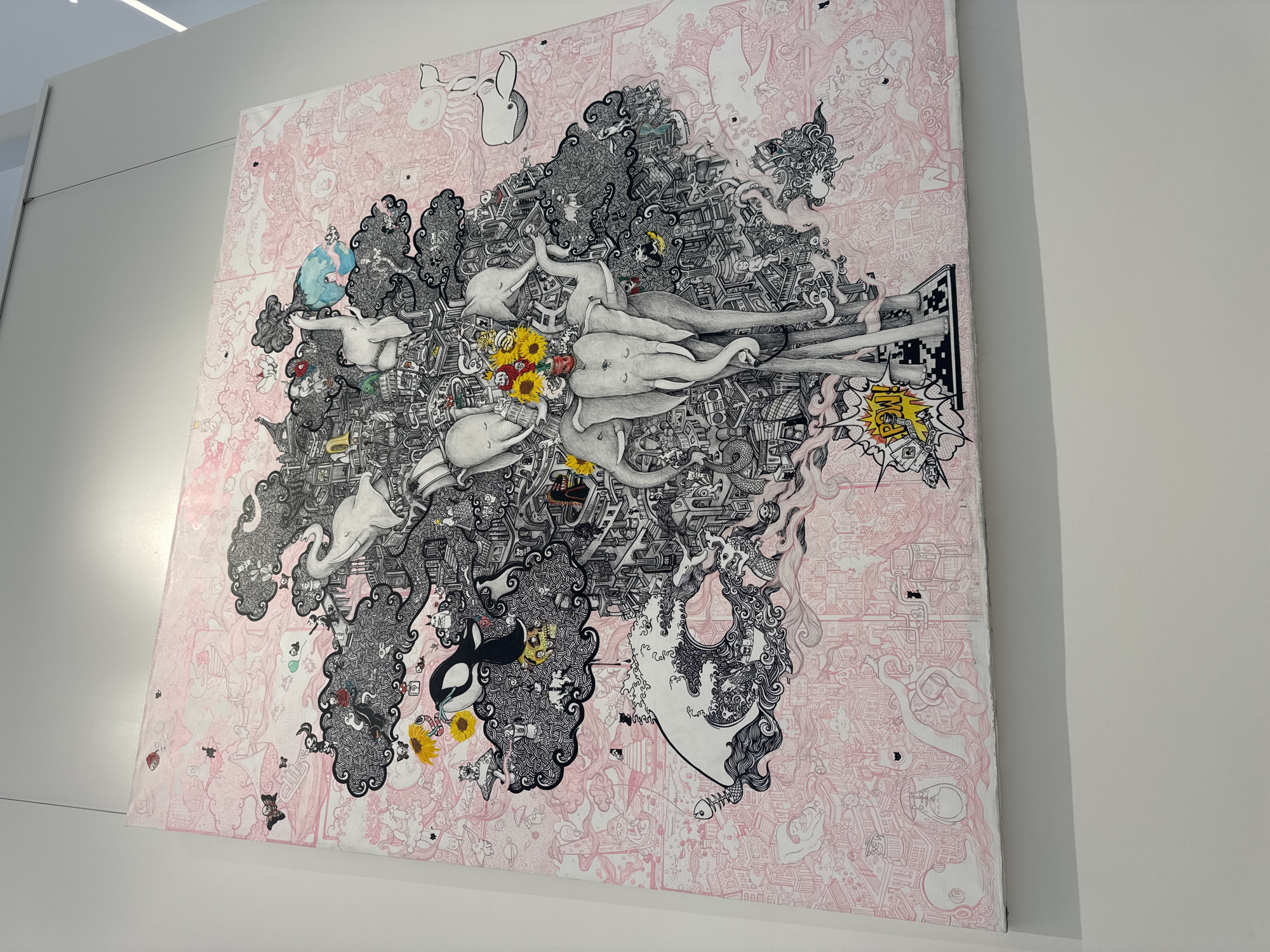 The image shows a large, square artwork hanging on a white wall. The artwork is densely detailed, primarily in black and white with touches of color. It depicts stylized elephants emerging from or embedded within a chaotic landscape of architectural elements, patterns, and other fantastical creatures. The background is a pale pink with delicate line drawings of buildings, people, and objects. The artwork is likely located indoors, possibly in a gallery or museum space, given the clinical white walls and professional presentation. There is no visible information confirming the location is Beverly Hills, United States of America.