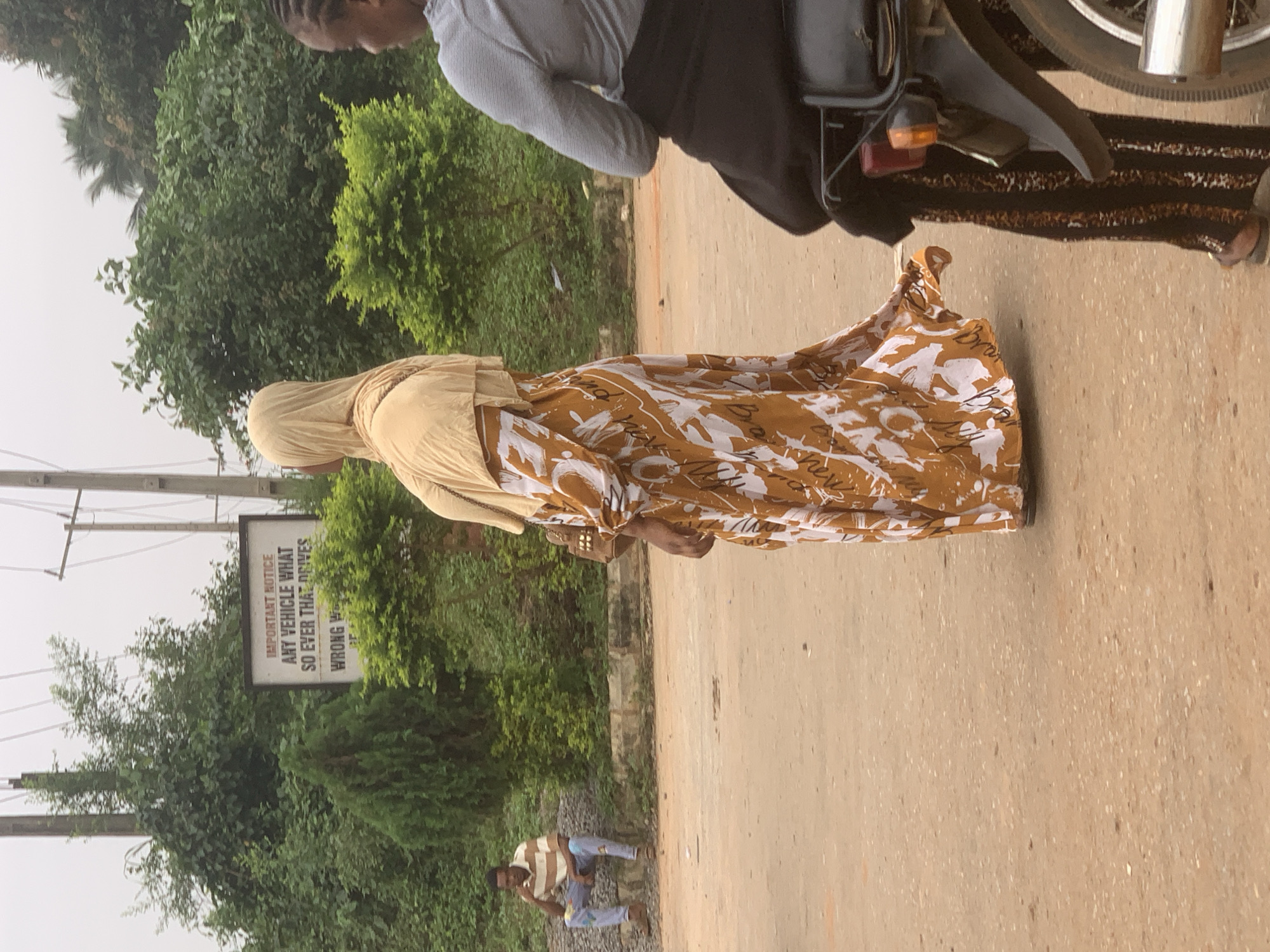 In Shagamu, Nigeria, a woman in a light yellow hijab and a patterned brown and white dress walks on a dirt road, her back to the camera. To her right, a person wearing a striped shirt and blue jeans sits on gravel. Behind them, lush green trees and a sign with text are visible. In the foreground, a motorcycle's rear wheel and taillight are partially visible, with a person on the motorcycle also seen from behind.