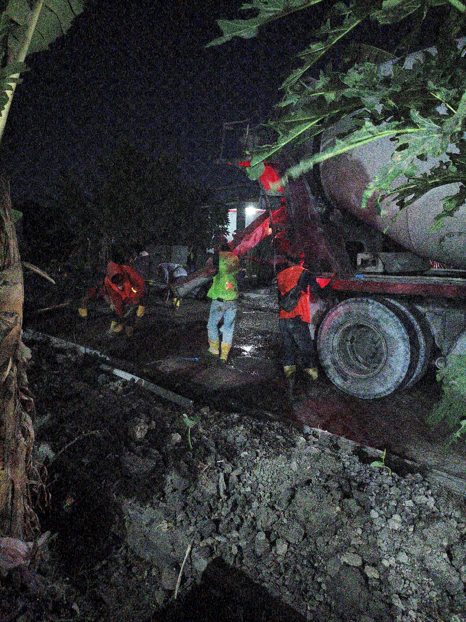 In the city of Gondangrejo, Indonesia, at night, workers are engaged in construction activities next to a cement mixer truck. The scene is dimly lit, with artificial lights illuminating the work area. Several individuals are visible, wearing high-visibility vests and work boots, indicating they are involved in manual labor. One person in the foreground is wearing a bright orange vest over a dark shirt and pants, and yellow boots, and appears to be overseeing the operation. Another worker in a green vest with reflective stripes and light blue pants is also present. The ground is uneven and appears to be a construction site with dirt and debris. The cement mixer truck is a prominent feature, with its large drum and wheels suggesting a substantial construction project is underway. The surrounding environment is dark, with foliage visible in the upper parts of the image, suggesting an outdoor setting. The overall impression is one of a nighttime construction operation in progress.