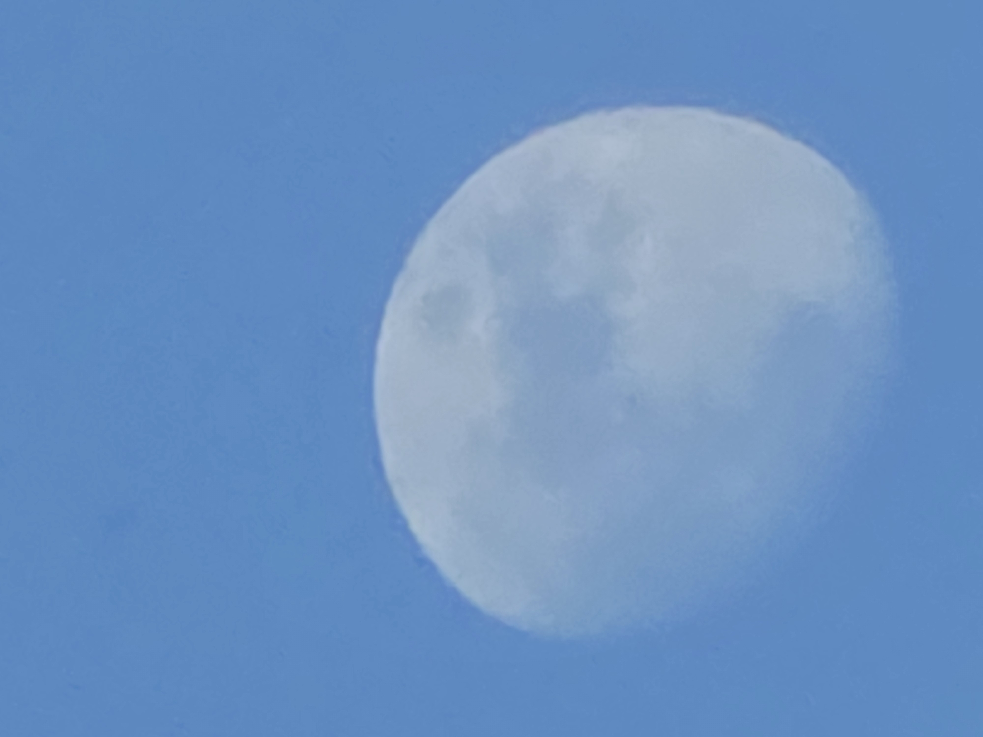 A daytime shot shows a waning gibbous moon against a pale blue sky. The moon is positioned on the right side of the frame, with a portion extending past the center of the image. Its surface has subtle variations in shading and texture. No people or objects are in the image, and the environment consists solely of the sky. There is no information available to ascertain the location is Varna, Bulgaria.