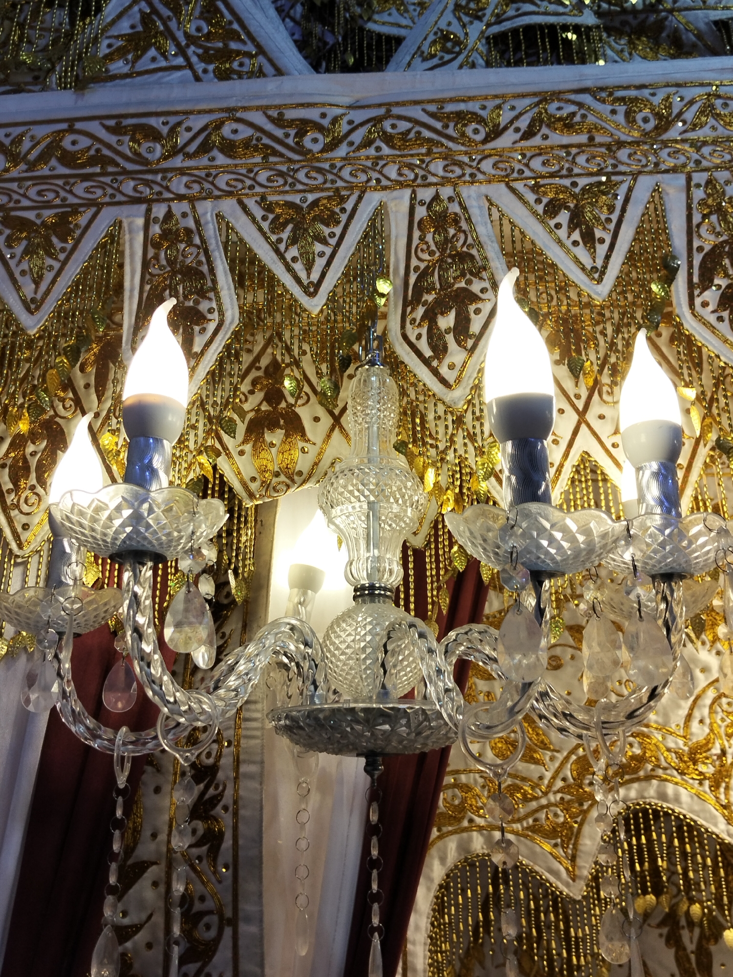 This image is a close-up shot of a crystal chandelier with four lit bulbs, set against a backdrop of ornate white and gold fabric with bead and sequin embellishments. The fabric has a repeating pattern of stylized leaves and swirling designs, with gold fringe hanging down. The chandelier itself features a central crystal stem and arms, adorned with teardrop-shaped crystal pendants. The lighting is warm, highlighting the textures and details of both the chandelier and the fabric. The exact location in Cottrueng, Indonesia is not discernible from the image.