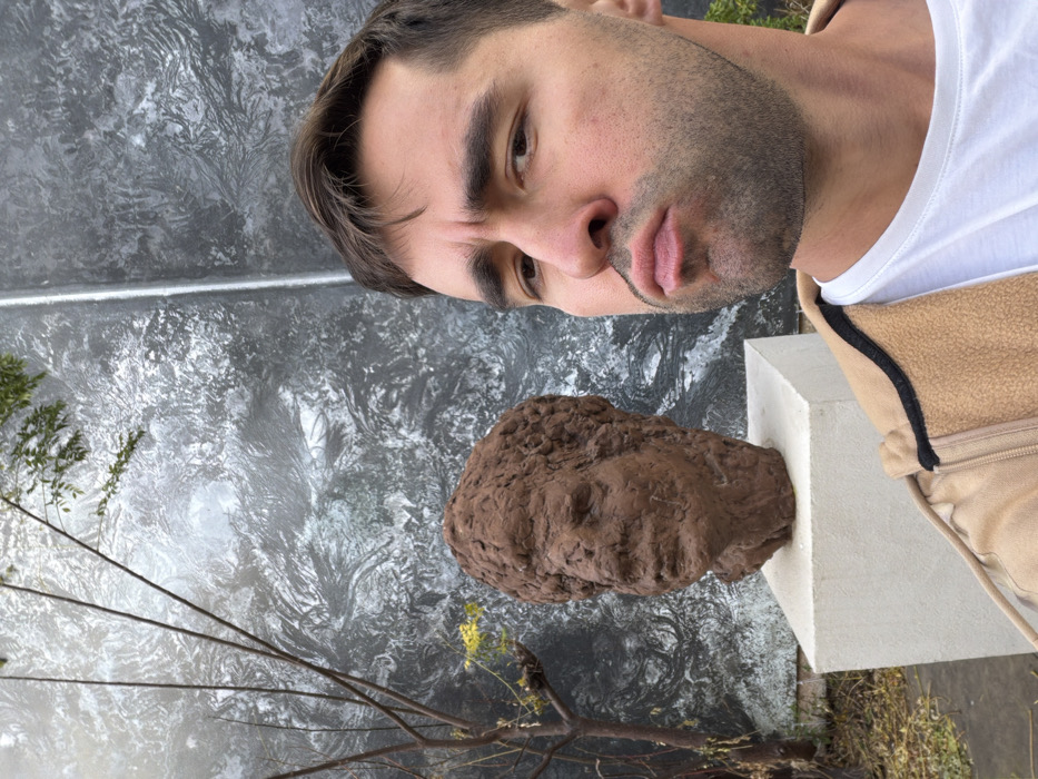 The image portrays an outdoor scene in Tel-Aviv, Israel, featuring a male subject and a sculpted bust. The man, positioned on the right and cropped from the chest up, is light-skinned with short dark hair, a trimmed mustache, and stubble. He wears a white t-shirt beneath a tan fleece jacket with black trim and exhibits a pouting expression while looking directly into the camera. To his left, slightly behind him, a reddish-brown, rough-textured bust depicting a head with dense, curly hair and discernible facial features rests on a light-colored, rectangular concrete pedestal. The background consists of a dark wall with an abstract, highly textured pattern resembling swirling frost or agitated liquid, featuring shades of grey and black. To the far left, behind the bust, sparse tree branches with minimal green foliage and yellow elements are visible against the textured wall, with dark ground cover at the base.