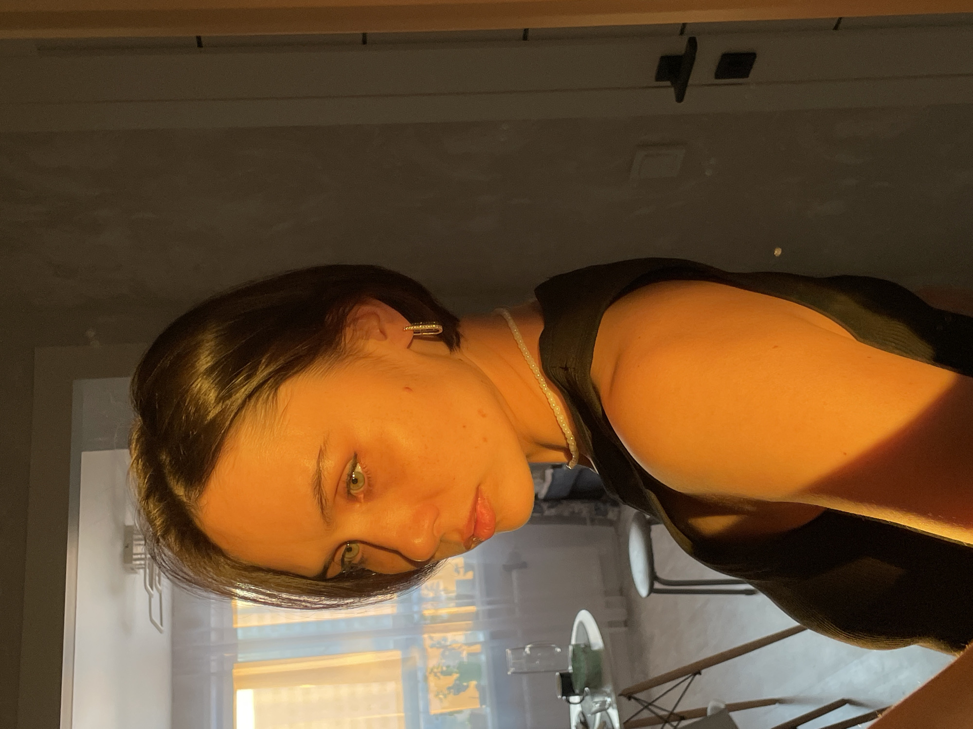 A medium shot depicts a young woman indoors, with the light appearing to be from a sunset or sunrise, as evidenced by the warm color temperature. She is positioned on the right side of the frame and appears to be looking downwards towards the lower left. The woman has short, dark hair and fair skin. She is wearing a dark top, and a pearl necklace is visible. A gold hoop earring is visible on her left ear. Her features are accentuated by the lighting, highlighting her eyes and lips.
The setting is an interior space, with sunlight streaming in through a window in the background. The interior has a minimalist aesthetic. The location in the city of Izhevsk, Russia is not possible to verify from the image.