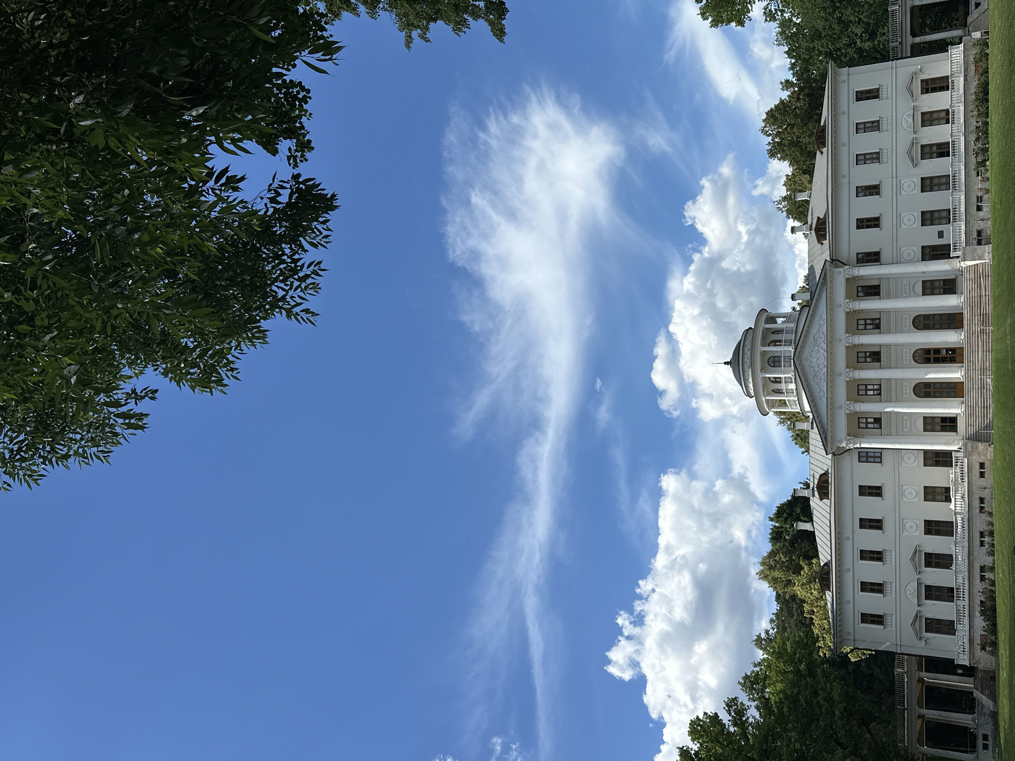 A white, classical-style building with a domed rotunda is viewed from a low angle, with the sky filling the majority of the frame. The building features multiple stories, windows with dark frames, and a portico with columns. Lush green trees frame the left side of the image, with their leaves appearing dark against the bright blue sky. Wispy white clouds are scattered across the sky, with a larger, more voluminous cloud formation on the right. The scene suggests a sunny day, likely in the afternoon, given the brightness of the sky and the shadows cast. The overall impression is one of architectural grandeur set within a natural, verdant landscape. No people are visible in the image. There is no visible text. The location context is Ostafievo, Russia.