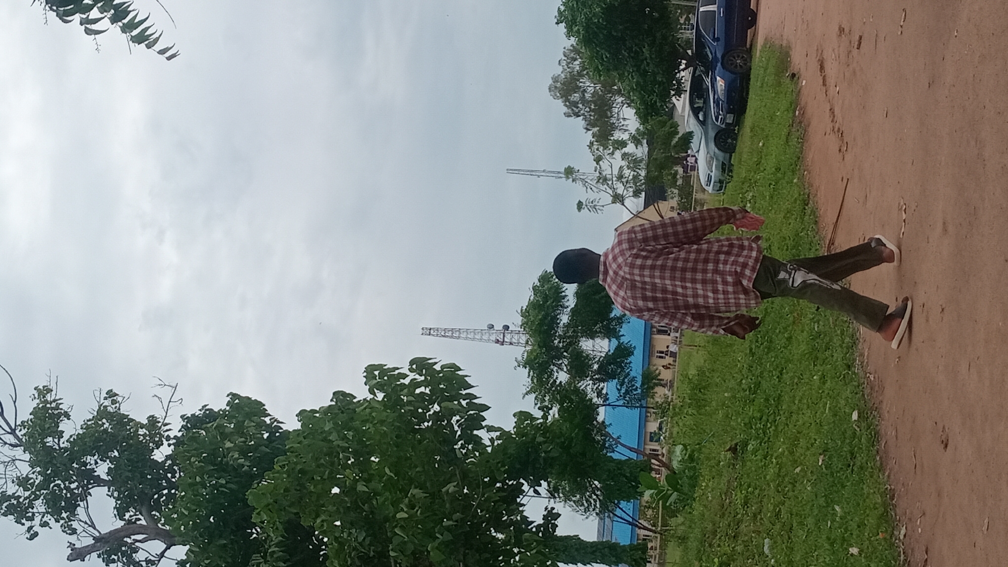 This media file captures a person walking along a dirt path outdoors. The subject is an adult, likely male, wearing a red and white plaid shirt and dark pants. They are walking away from the camera, with their right arm slightly extended. Their gait suggests they are in motion, possibly walking at a moderate pace.
The setting appears to be a semi-rural or suburban area, with a mix of natural and man-made elements. Lush green grass borders the dirt path, and several trees with dense foliage are visible in the foreground and background. In the distance, a tall telecommunications tower pierces the overcast sky. A blue-roofed building is also present, with some people gathered in front of it. Two cars are parked on the left side of the frame, one a dark blue sedan and the other a lighter colored vehicle.
The weather seems to be cloudy or overcast, as indicated by the uniformly gray sky. This suggests it might be daytime, but the exact time is unclear. There are no visible signs or text within the frame. The overall mood of the image is neutral, focusing on the simple act of a person walking through an everyday environment. The scene conveys a sense of movement and a typical outdoor day.