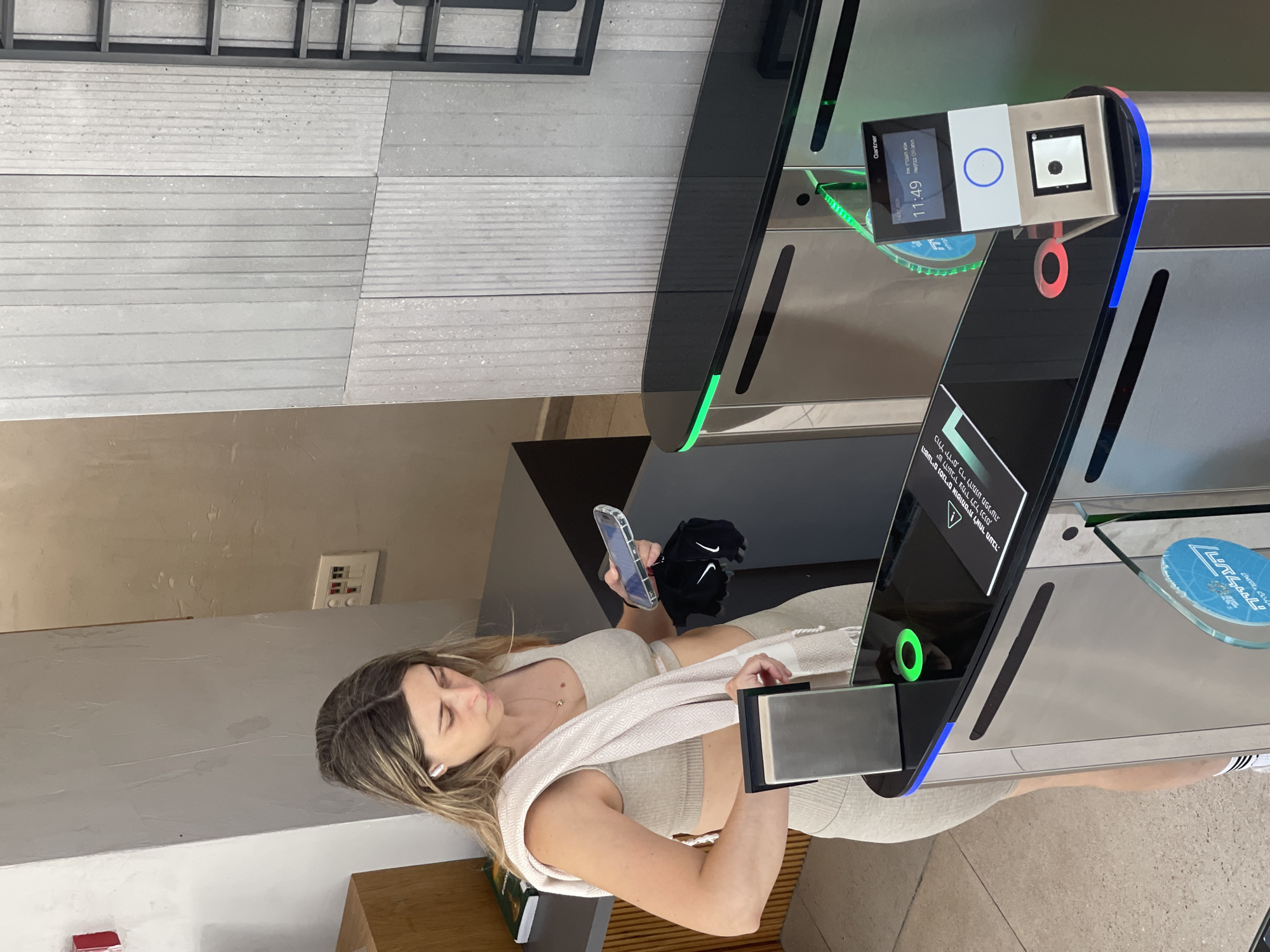A woman with blonde hair, wearing a beige athletic outfit and white earbuds, stands in front of a modern turnstile in Tel Aviv, Israel. She is holding a smartphone in her right hand and a white towel is draped over her left shoulder. The turnstile has a black glass top with green and blue LED lighting and a digital display showing the time as "11:49" and some text in Hebrew. A small scanner is visible at the bottom of the display. The environment appears to be an indoor public space, possibly a gym or office building entrance, with a gray tiled wall in the background.