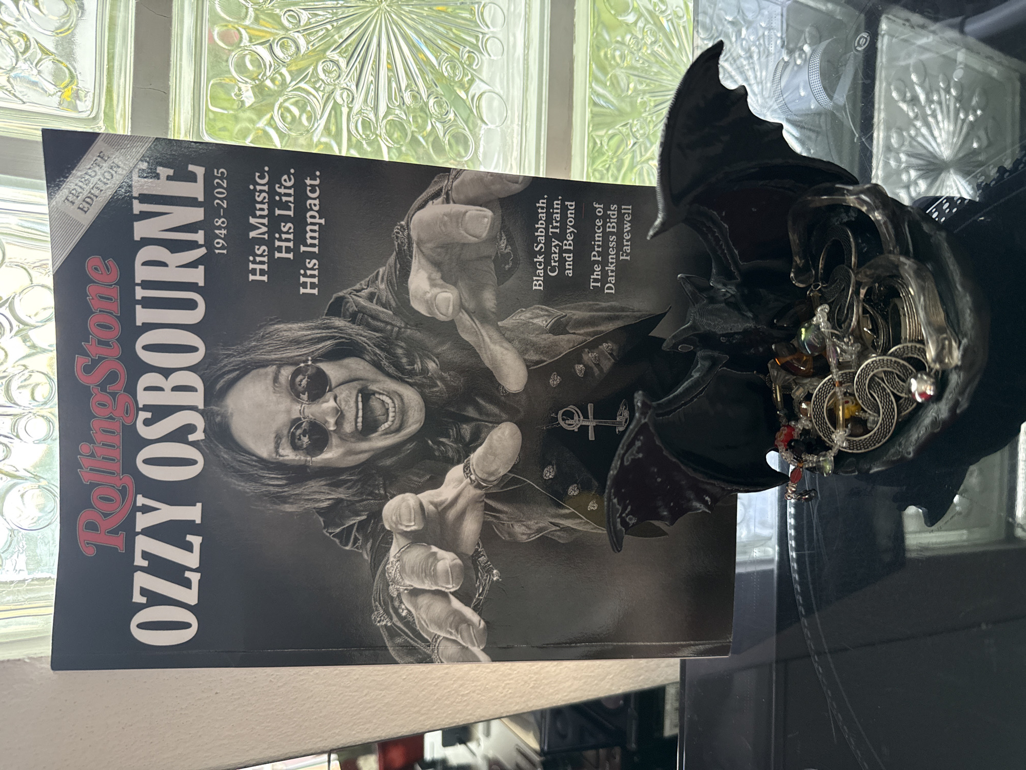 A close-up, eye-level shot shows the cover of a Rolling Stone magazine featuring Ozzy Osbourne. The magazine is titled "OZZY OSBOURNE 1948-2025" and has a subtitle that reads "His Music. His Life. His Impact." A small banner at the top right corner of the magazine reads "TRIBUTE EDITION." Osbourne is pictured in black and white, with a wide-mouthed expression and wearing round sunglasses. He is wearing multiple rings on his fingers and a pendant necklace. Below the magazine, a dark, reflective surface holds a decorative black bat-shaped bowl filled with assorted jewelry, including rings and necklaces. The background is out of focus and appears to be a window with patterned glass, possibly illuminated by natural light. The scene does not appear to be located in the city of Ko Samui District, Thailand.