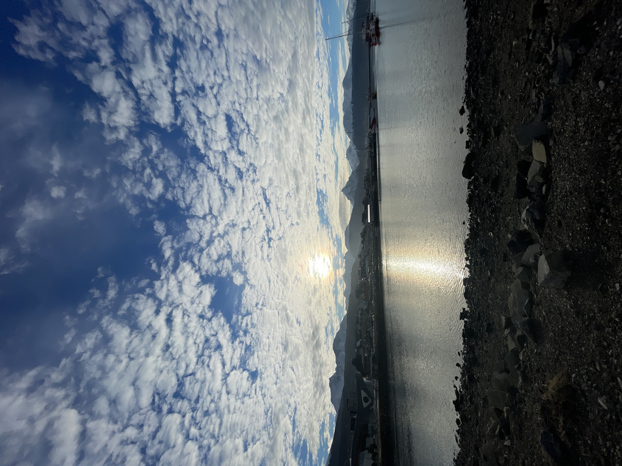 The image depicts a harbor scene, likely Ushuaia, Argentina, under a sky filled with altocumulus clouds. Sunlight is diffused by the clouds, yet a distinct reflection of the sun shines off the water's surface. Mountains loom in the background, with their peaks partially covered in snow. A few buildings and industrial structures, possibly related to shipping or port activities, are visible along the shoreline. The immediate foreground consists of a rocky shoreline. There are boats visible in the harbour.