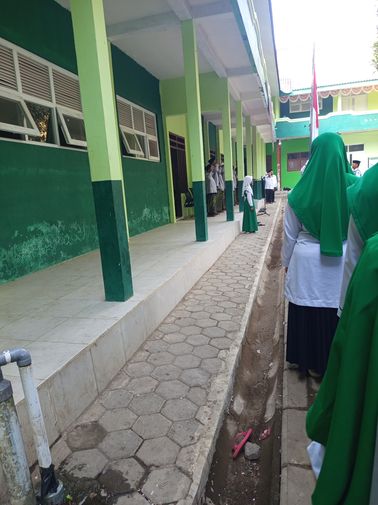 In Lumutan, Indonesia, this image captures an outdoor scene along a covered walkway of a green building. The walkway is paved with hexagonal concrete tiles and has a drainage ditch running alongside it. Several individuals, predominantly young people, are present, many wearing green headscarves or hijabs and dark skirts or trousers, suggesting they are students. Some are standing in lines, facing towards a flagpole that is visible in the background with the Indonesian flag. Others are walking or standing on the walkway. The building has a green exterior with rows of white-framed windows that appear to be louvered. The pillars supporting the walkway roof are painted green and a darker green. In the foreground, a water pipe and faucet are visible next to a tiled area. A single pink flip-flop and a stone lie in the drainage ditch. The lighting suggests it is daytime, possibly overcast.