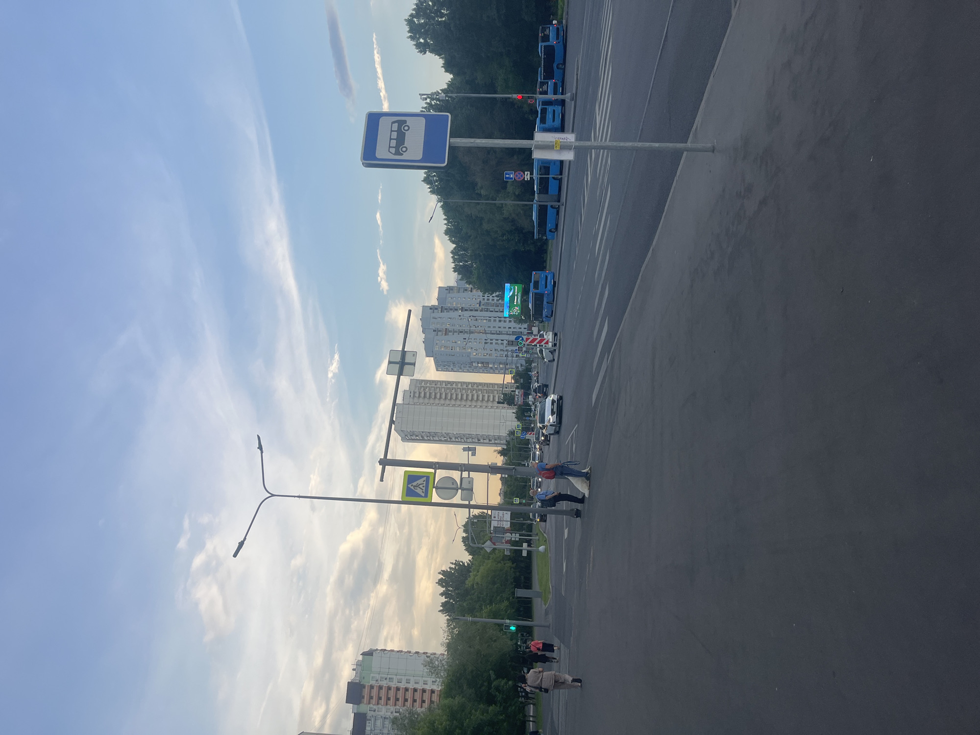 The image depicts a street scene in Moscow, Russia, with multiple tall apartment buildings in the background. On the left side of the street, several pedestrians are walking on the sidewalk. A traffic light is visible, showing a green signal. The street itself has a crosswalk and several cars are driving on it. On the right side of the street, a bus stop sign is prominently displayed, with several blue buses lined up at the stop. The sky is partly cloudy, with a mix of blue sky and white clouds. The overall impression is that of a busy urban environment in the late afternoon or early evening.