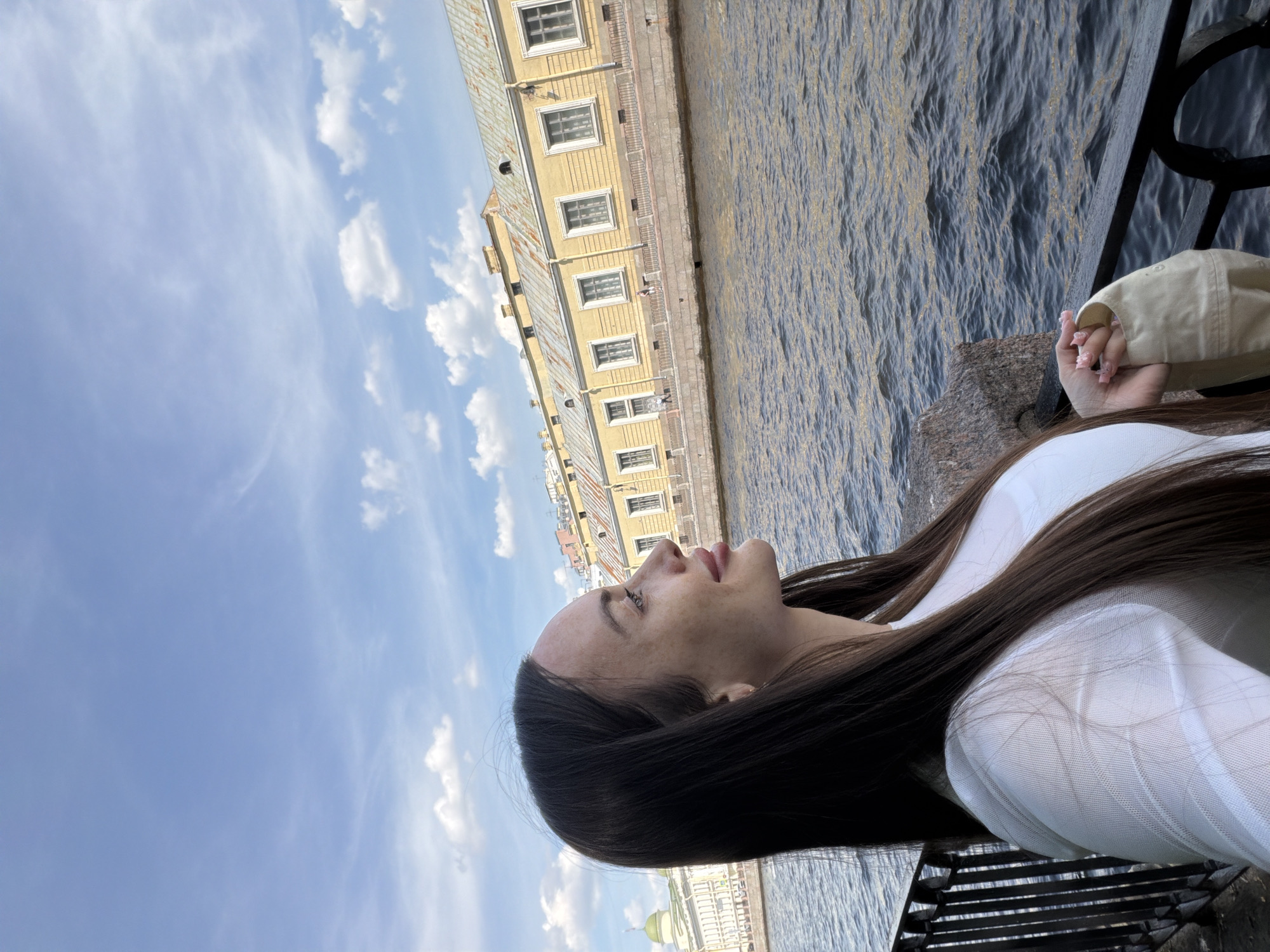 A young woman stands on a waterfront promenade in Saint Petersburg, Russia, facing left in profile. She has long, dark brown hair and is looking up and to her left. She is wearing a white top with a patterned texture and is holding a beige jacket or bag in her hands. Behind her is a canal with dark, choppy water. Across the canal, a long, yellow building with a slightly rusted roof and numerous windows lines the embankment. The sky above is blue with scattered white clouds. In the distance, a green dome is visible, suggesting the presence of a church or other significant architectural structure.