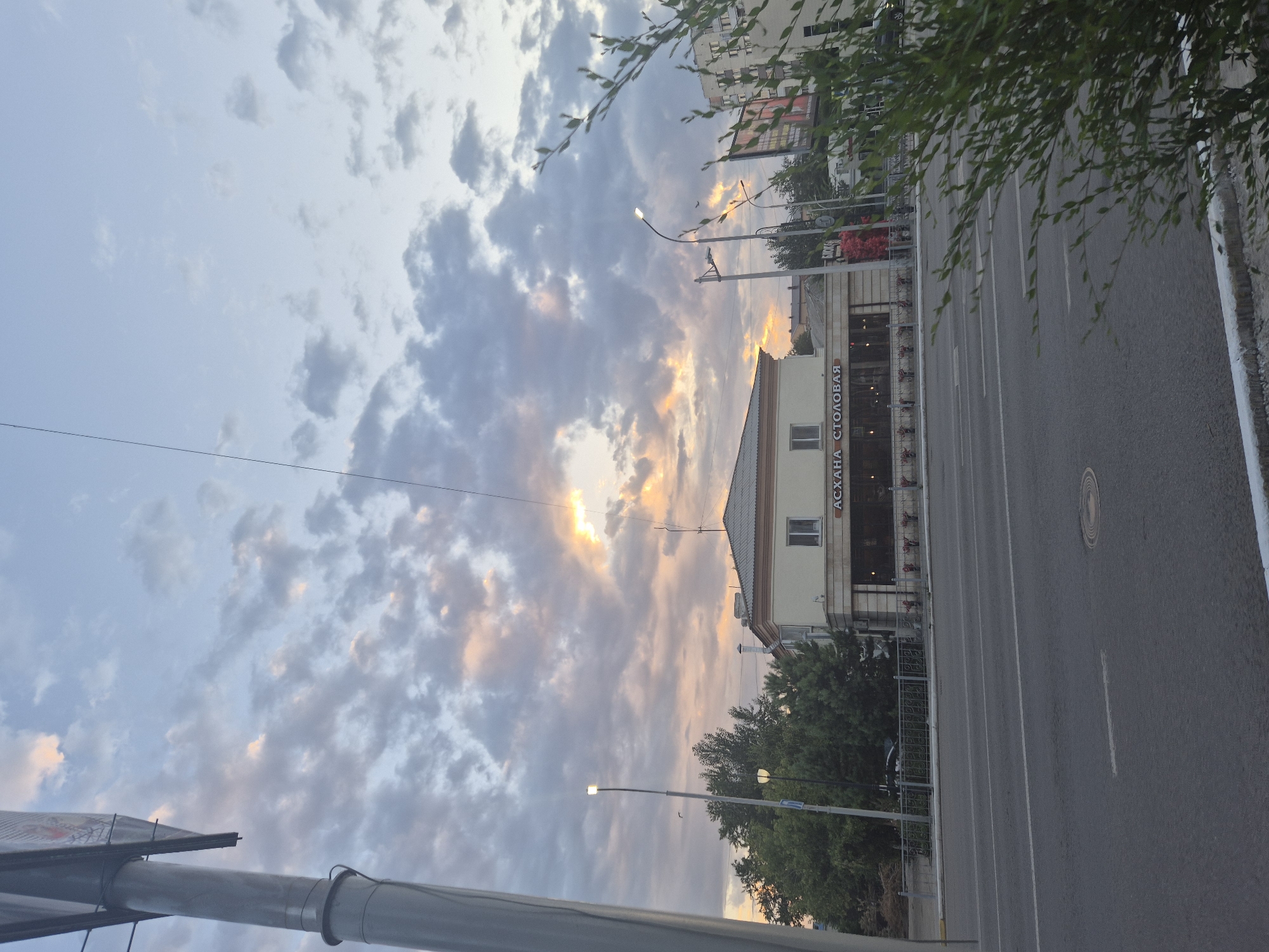 The image depicts a street scene in Kokshetau, Kazakhstan, during a sunset. The sky is filled with dramatic clouds, with rays of sunlight breaking through, illuminating the scene with warm hues. In the foreground, a grey asphalt road with white lane markings is visible. To the right, lush green foliage partially obscures the view.
Dominating the mid-ground is a two-story building with a light-colored facade. Above the ground floor windows, in Cyrillic script, the words "АСХАНА СТОЛОВАЯ" are clearly visible, indicating it is a canteen or dining hall. The building has a traditional architectural style with a pitched roof. Streetlights are positioned on the left side of the road, casting light. Trees line the street behind the building. In the background, other buildings and structures are faintly visible, including a large billboard. The overall impression is one of a serene evening in an urban environment.