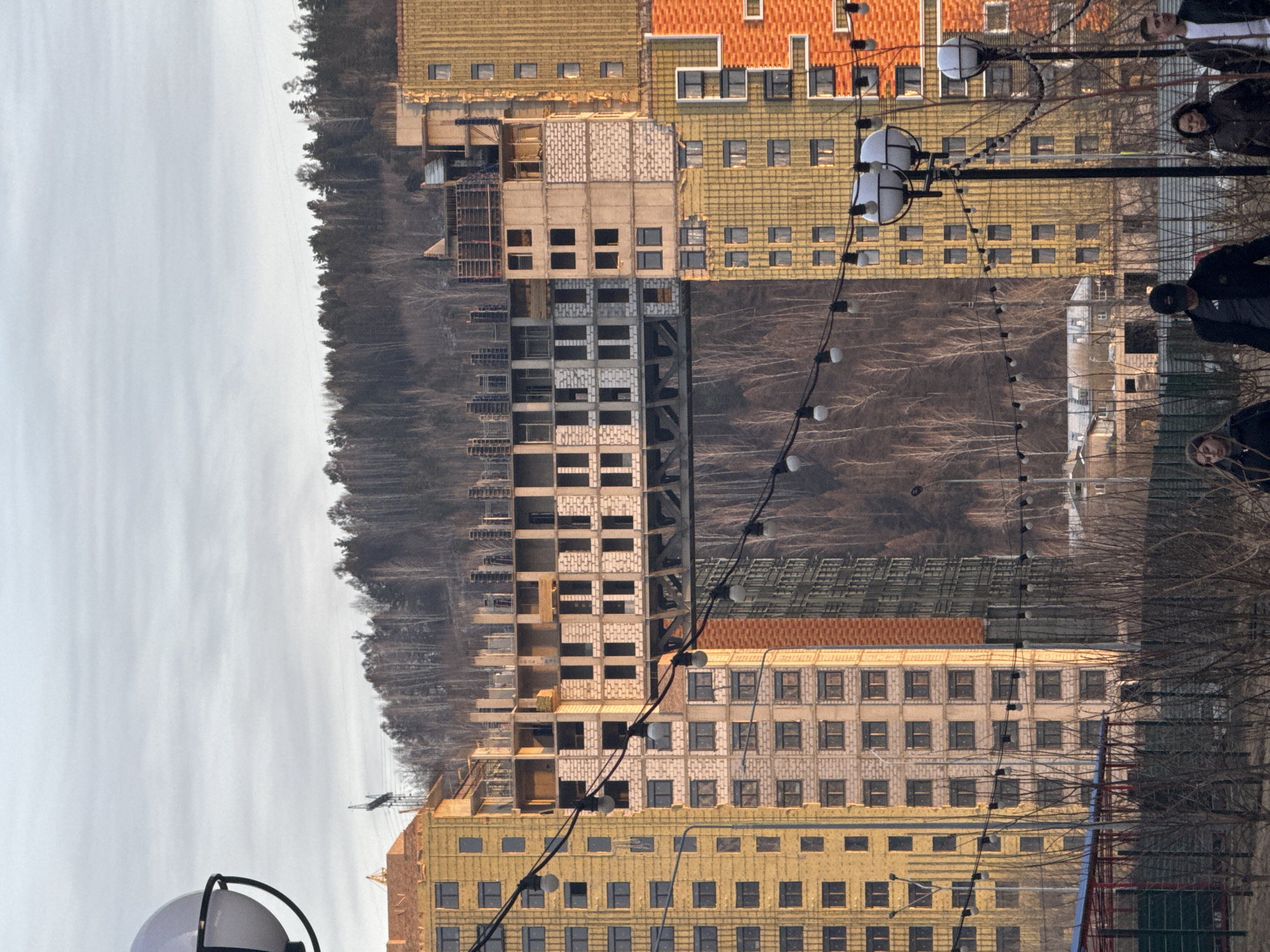 The image shows a cityscape in Krasnoyarsk, Russia, featuring a mix of completed and under-construction buildings. The buildings have facades in varying colors, including yellow, orange, and grey. Some buildings are framed with scaffolding. A forested area rises along the background. Street lights with spherical lamps are present. There are a few individuals visible in the right edge of the frame, wearing dark clothing. Overhead string lights also run across the foreground. The sky is overcast.