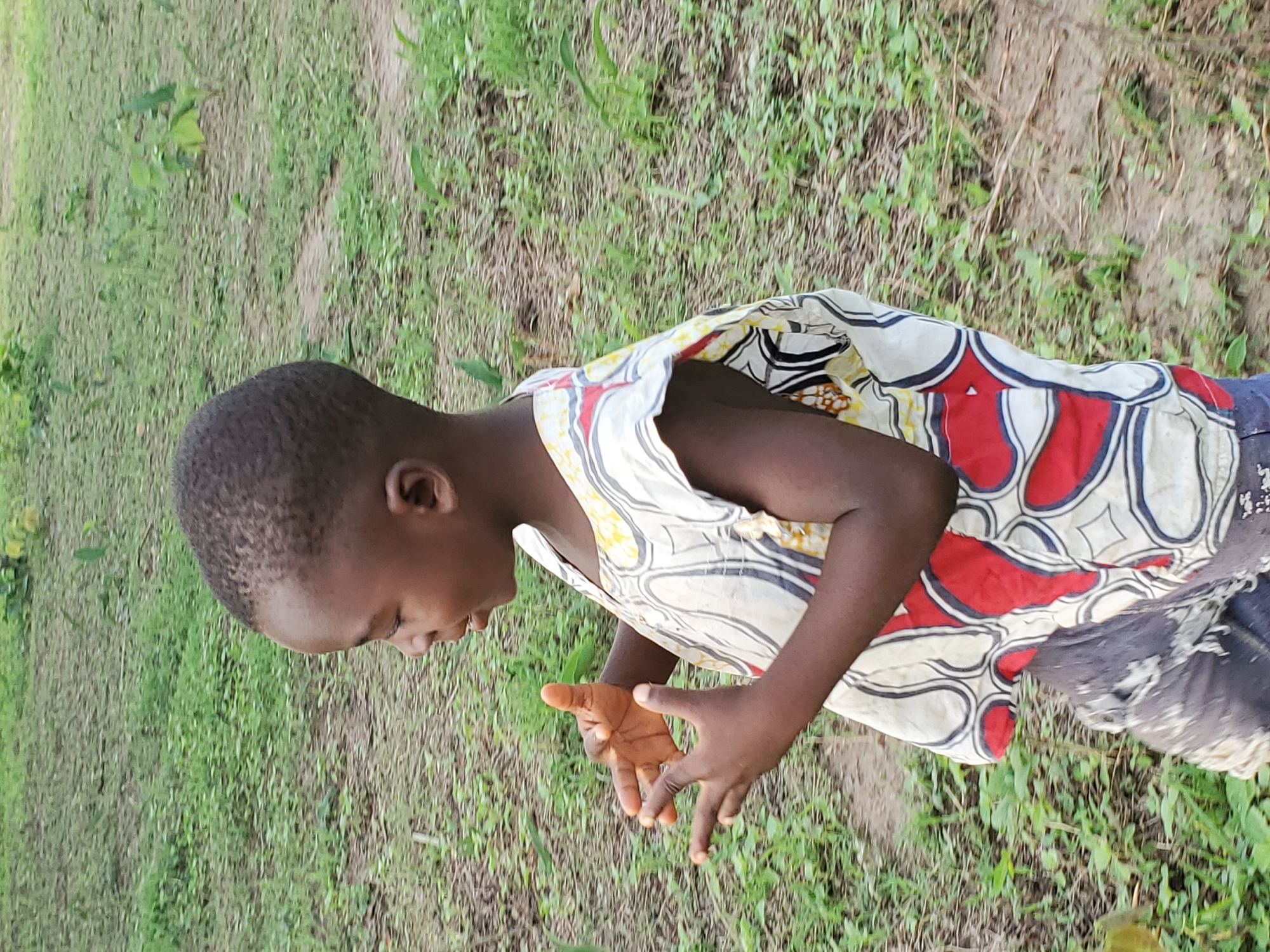 A young boy with dark skin is standing in a grassy area. He is looking down and gesturing with his hands. The boy is wearing a sleeveless shirt with a pattern of white, red, black, and yellow. The background is green, with grass and some small plants visible. The location is Mararaba, Nigeria.