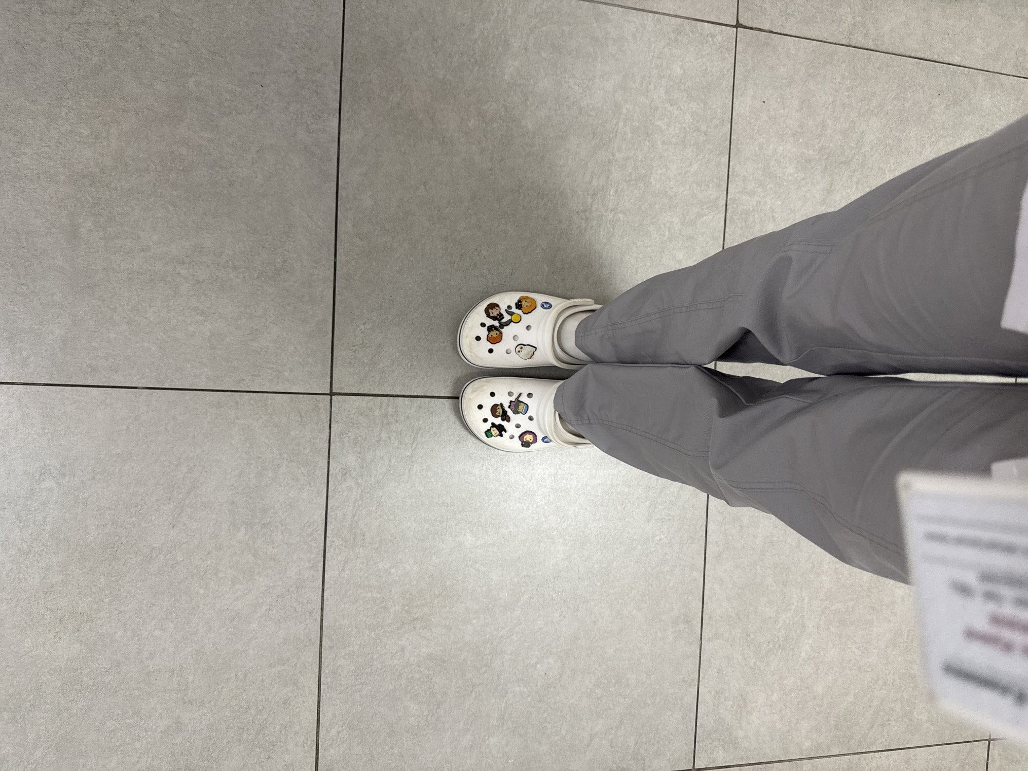 The image shows a person's feet and lower legs on a gray tiled floor. The person is wearing gray pants and white Crocs adorned with cartoon character charms. The location is unknown and there is no indication of it being in Moscow, Russia.