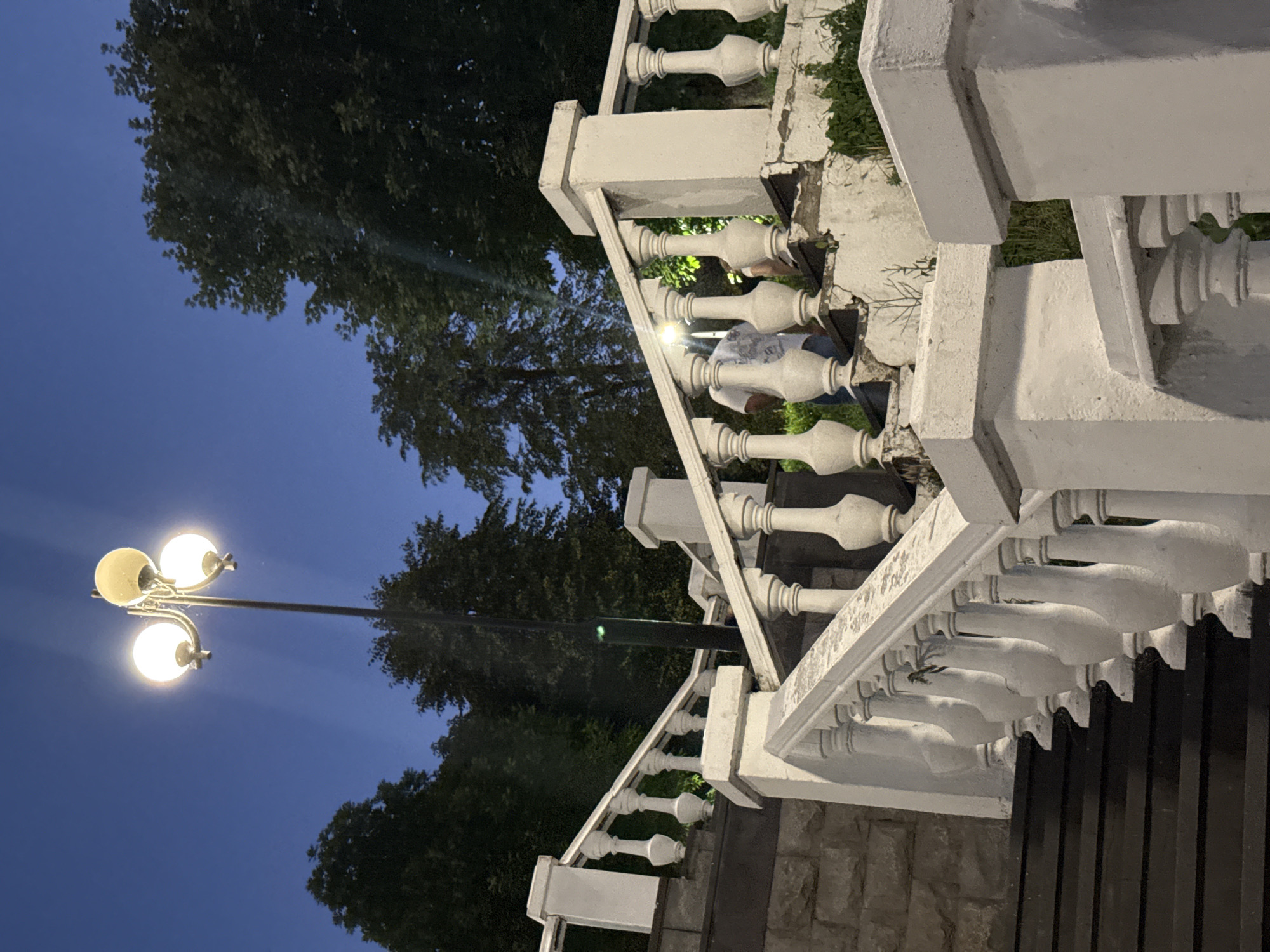 The image depicts an outdoor stairway at dusk in Endirei, Russia. A multi-globe lamppost illuminates the scene, casting rays into the dark blue sky. The stairway is flanked by white balustrades with decorative pillars. Dark wooden steps are visible on the left. In the background, trees form a dense canopy against the fading light. Two individuals are present on the stairs, one in a white t-shirt facing away from the camera, and another figure partially visible further up the stairs. The overall atmosphere is calm and serene, with the artificial light from the lamppost contrasting with the natural twilight.