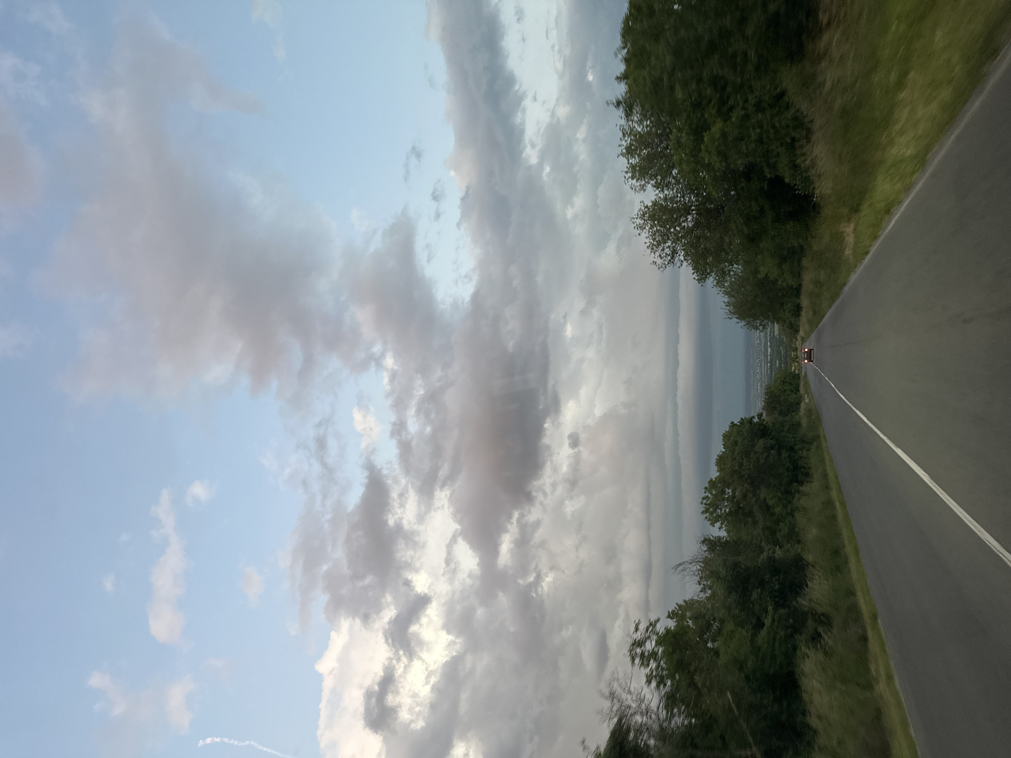 The image shows a road receding into the distance, flanked by trees on either side. The sky is filled with clouds, some light and others darker. At the end of the road, what appears to be a city can be seen in the distance. A car with its lights on is visible in the distance.
The location is Pryaporets, Bulgaria.