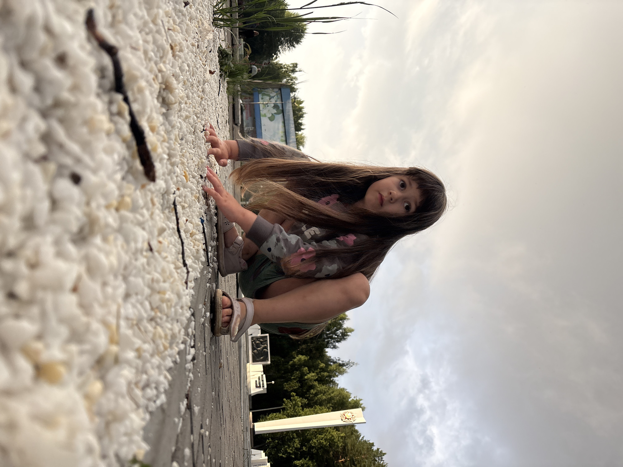 A young girl is crouching low on a paved area covered in white gravel. She is looking directly at the camera with a serious expression. Her long, dark hair falls around her face. She is wearing a grey long-sleeved shirt with a floral pattern and teal shorts. Her feet are clad in light-colored sandals. The foreground is dominated by the gravel and a few scattered twigs. In the background, a white obelisk with a circular emblem is visible, along with trees and other structures, suggesting a public park or memorial site. The sky above is overcast with diffused light. The scene appears to be located in Chaikovsky, Russia, likely at a memorial complex or park area.