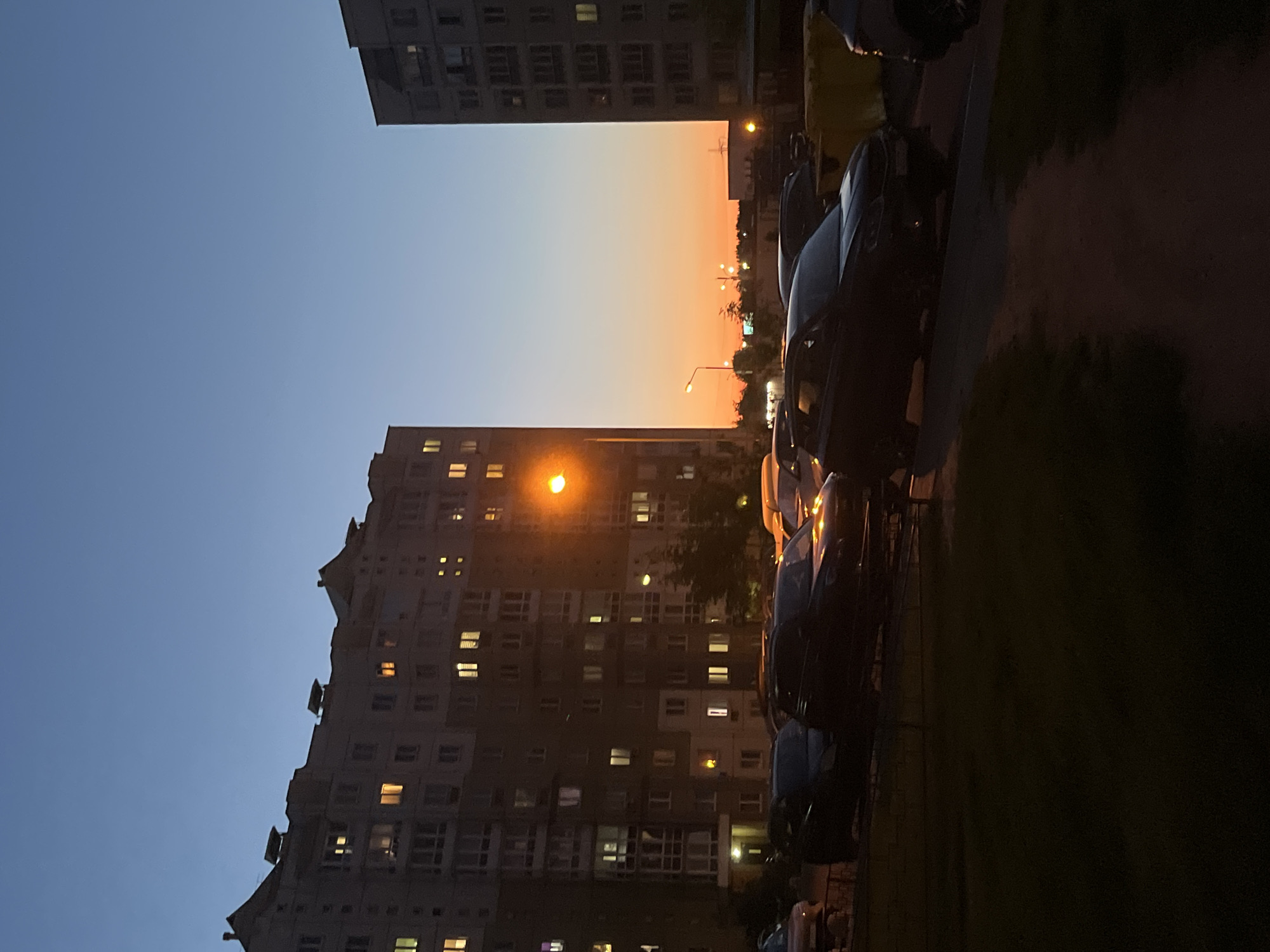 The image, correctly oriented (rotated 90 degrees clockwise), captures an urban residential scene at twilight in Minsk, Belarus. The sky transitions from a clear, deep blue at the top and left to a vibrant orange and pink gradient towards the horizon on the right, indicative of either sunset or sunrise.
On the left, a large, multi-story apartment building stands prominently, its dark facade punctuated by numerous lit windows, suggesting inhabitants are home. This building features a distinctive, somewhat jagged or stepped roofline. Another section of a similar apartment building is partially visible in the upper right quadrant.
Along the right side of the image, a street or parking area is lined with several cars, appearing as dark silhouettes against the ambient light. Streetlights are illuminated, casting an orange glow that contributes to the warm tones in the lower and right parts of the scene. Some dark foliage, likely trees or bushes, can be discerned near the street and cars.
The scene conveys a quiet, peaceful urban evening atmosphere. No people are visible, and there is no discernible text within the image. The architecture of the buildings is characteristic of residential areas found in many post-Soviet cities.