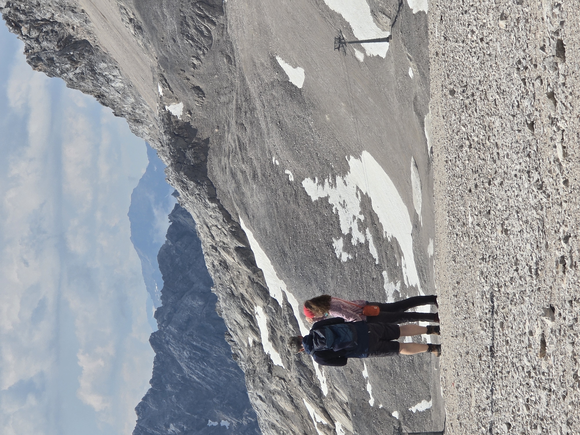 The image shows a man and a woman standing on a rocky, sparsely vegetated mountain slope with patches of snow. The man, wearing a backpack and shorts, stands to the left, while the woman, wearing a pink jacket and red hat, stands to his right. They are looking out at the mountainous landscape, which includes rugged peaks, a ski lift tower, and a cloudy sky. The scene is likely from the Zugspitze, Germany's highest peak, located near Garmisch-Partenkirchen. The overall impression is one of a dramatic, high-altitude environment.