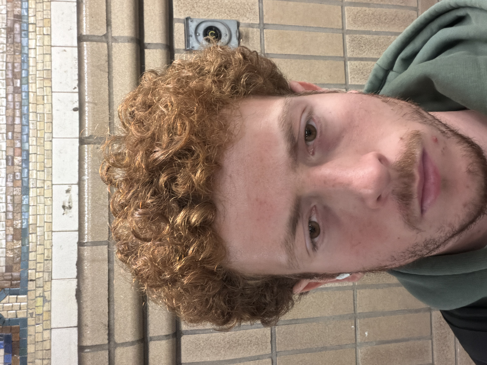 A close-up, eye-level photograph depicts a fair-skinned male with curly auburn hair, a trimmed auburn beard, and green eyes. He is wearing a dark green hooded sweatshirt. In his left ear, there is a white earbud-type device. The subject appears to be positioned against a tiled wall. The tiles behind him are a neutral tan, arranged in a brick pattern. Along the left edge of the frame, there are decorative mosaic tiles in a vertical strip of light beige, tan, white, and blue. An electrical outlet is visible at the top left of the man's head, indicating that the subject might be in a transit station or a commercial corridor in NYC.